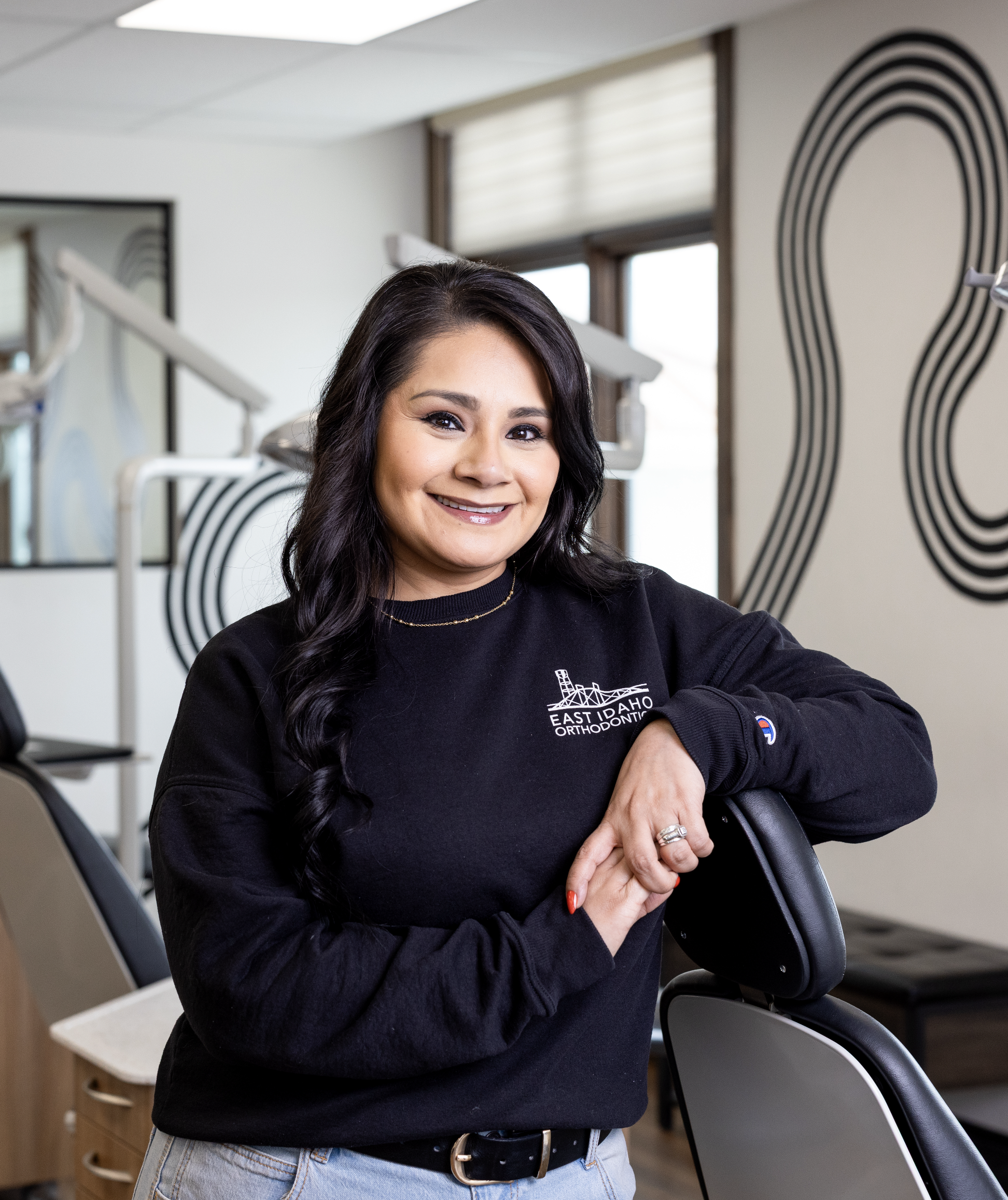 A woman smiling in a dental office, wearing a black sweatshirt with the logo East Idaho Orthodontic, leaning on a dental chair.