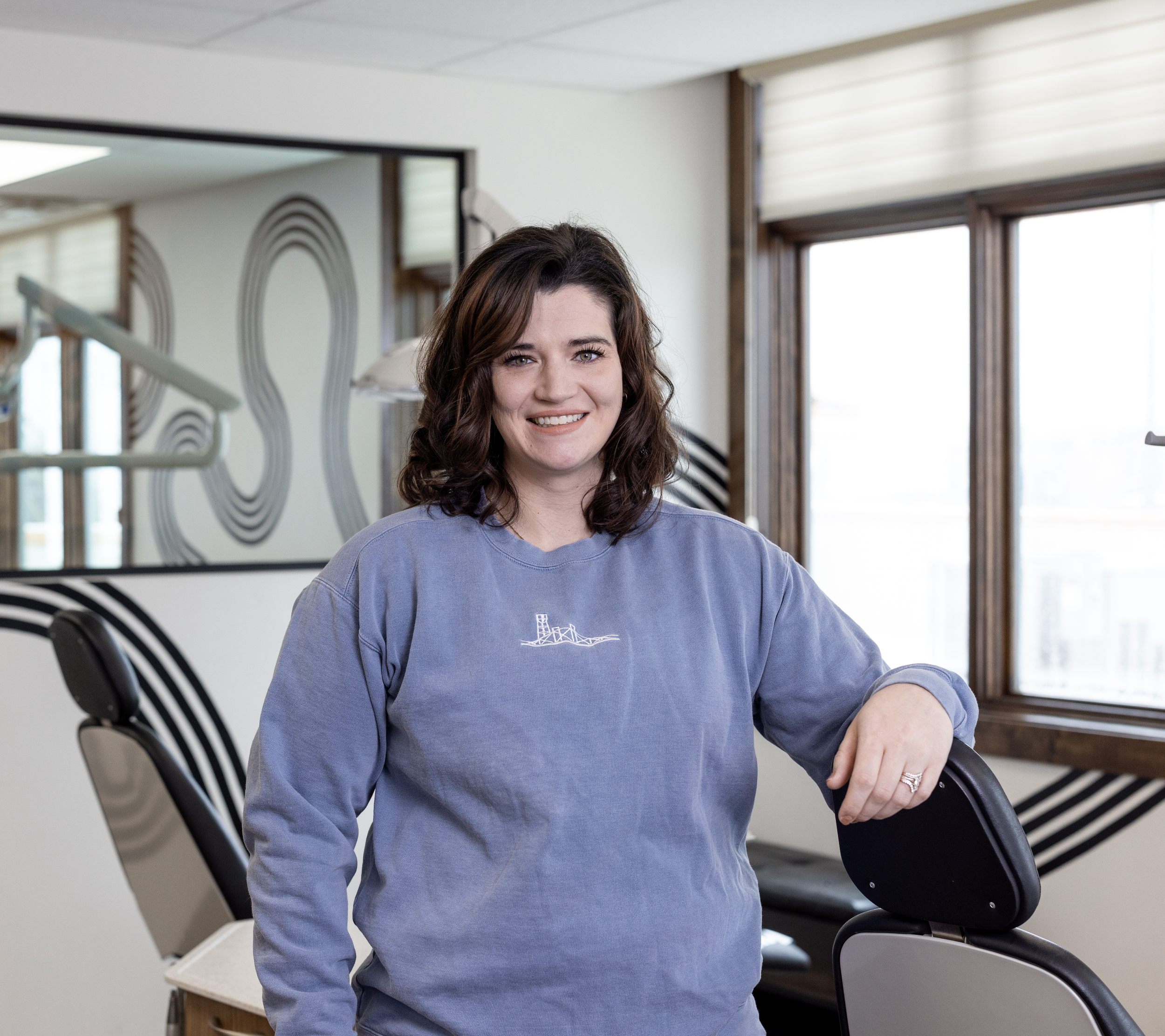 A woman with shoulder-length dark brown curly hair smiling, standing in a modern indoor space with large windows, a black chair, and a wall mirror in the background.