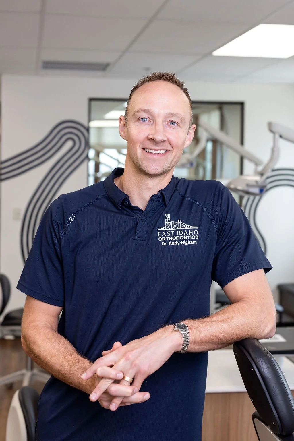 Dr. Higham in a navy blue polo shirt with the logo and text 'East Idaho Orthodontics Dr. Andy Higham' stands in an office with modern decor and dental equipment in the background.