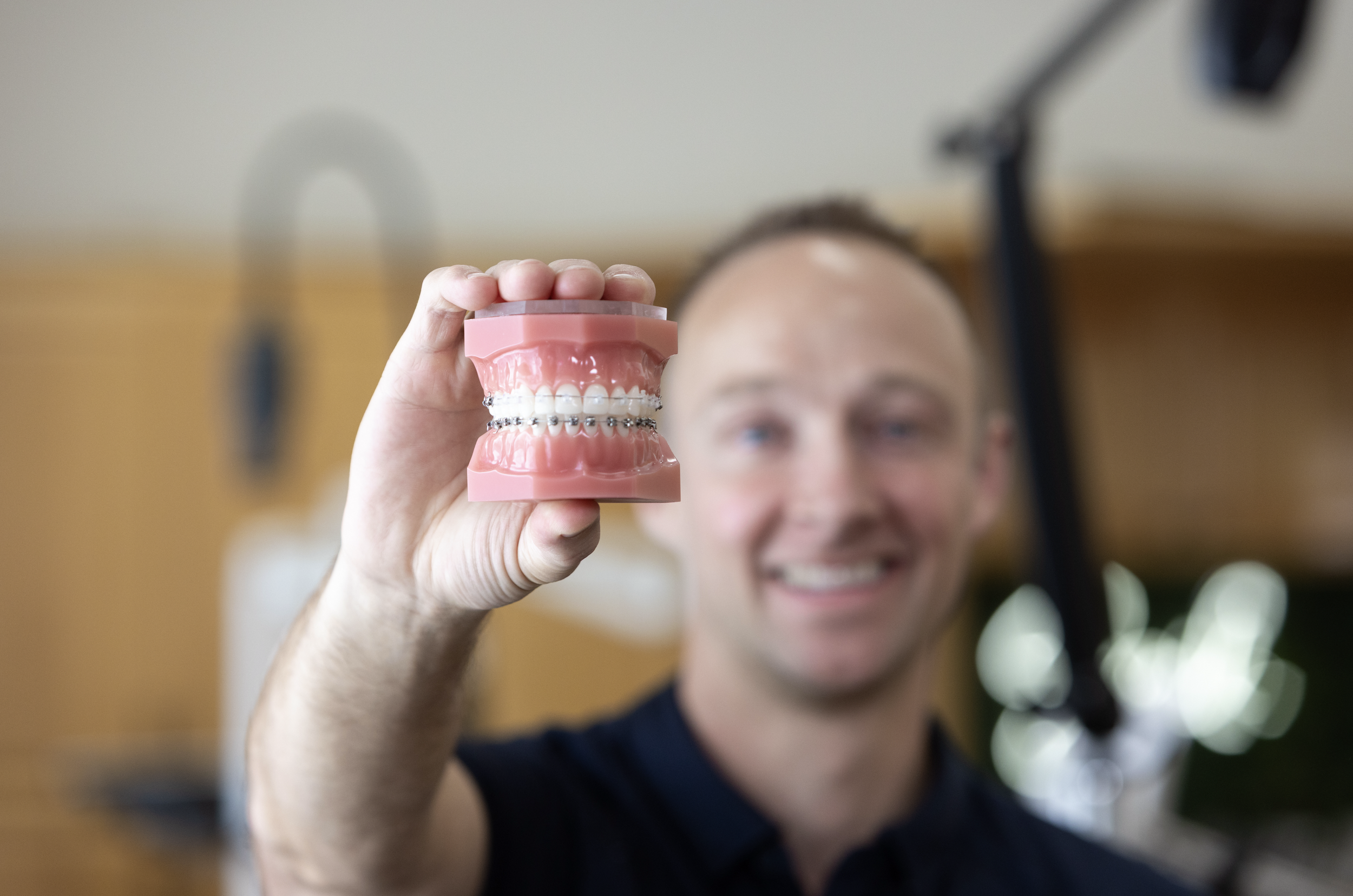 A smiling man holding a dental model with braces, with dental equipment in the background.