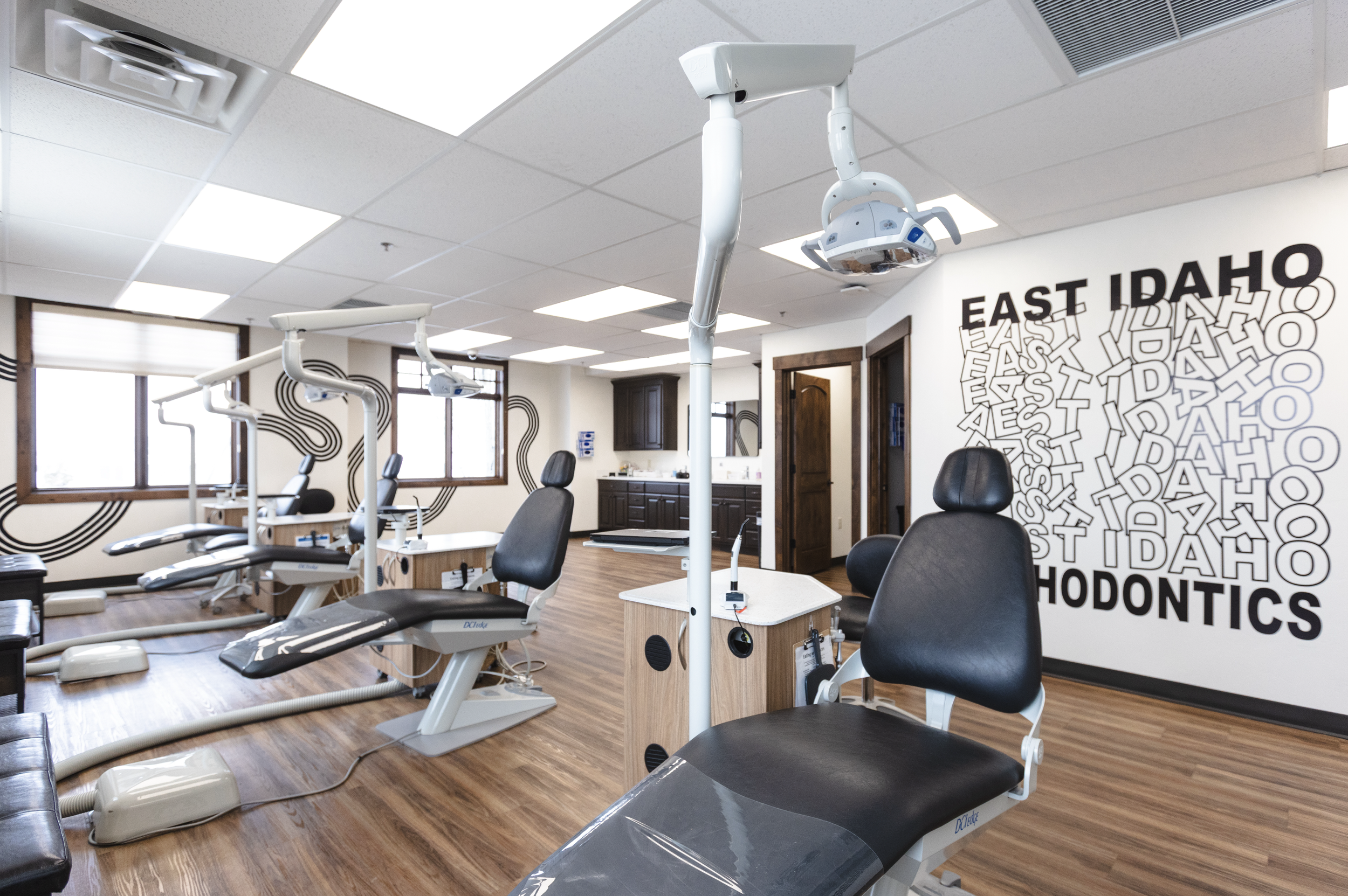 Dental examination chairs in a modern clinic with wooden floors, large windows, and a wall decorated with 'EAST IDAHO DENTAL' in bold letters and an abstract pattern.