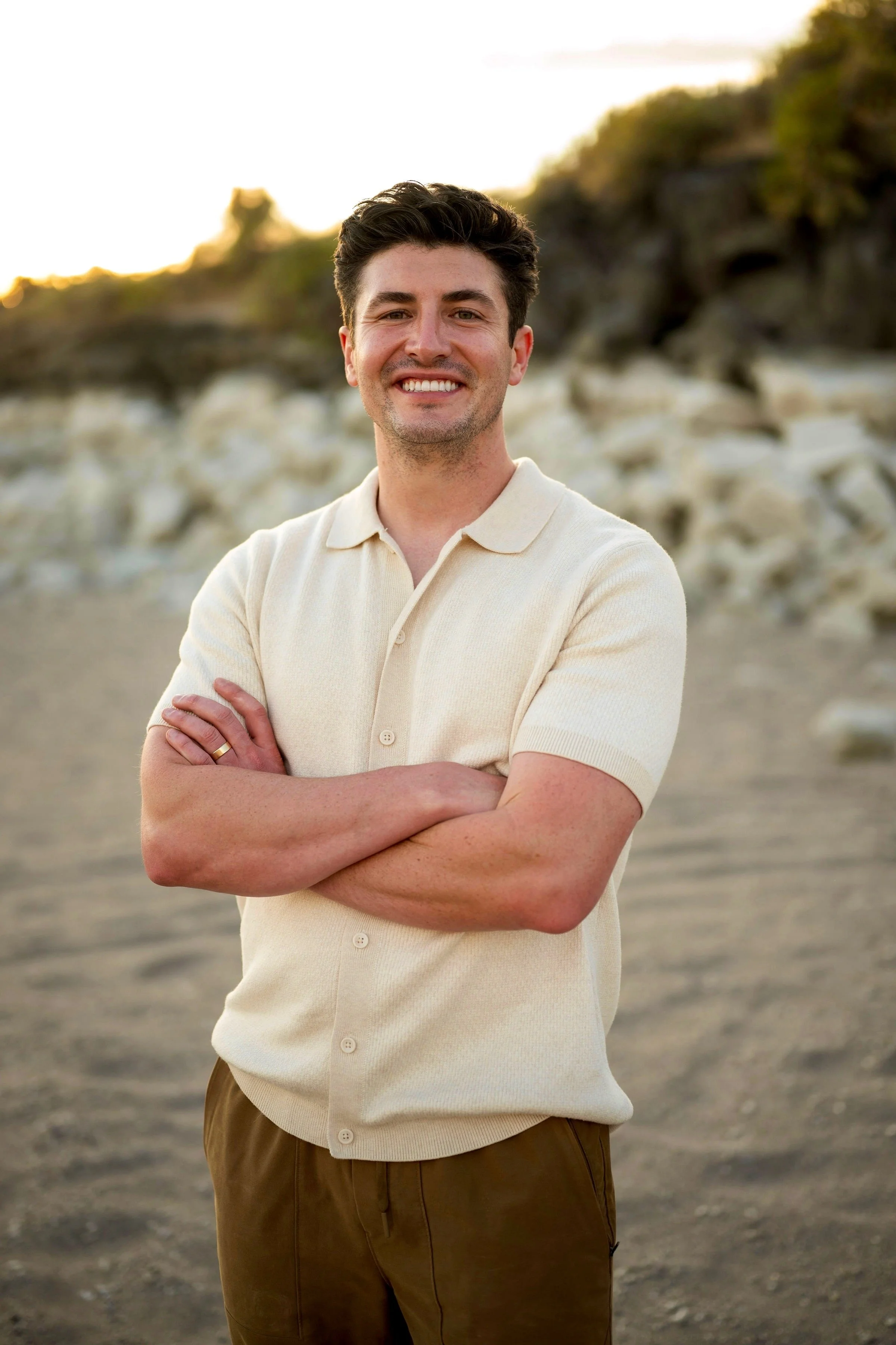 A man with dark hair and a smile, wearing a light beige short-sleeved shirt and brown pants, standing outdoors on a rocky, sandy area with a blurred natural landscape background during sunset.