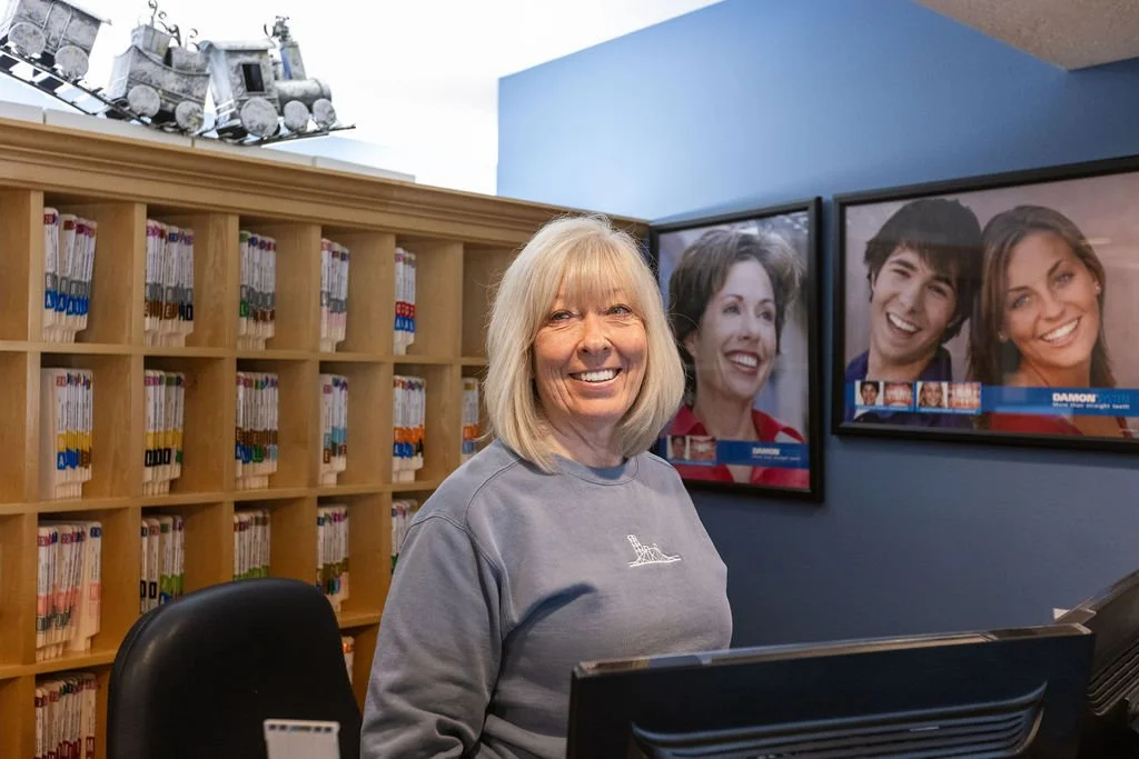 A woman with blonde hair smiling at a service desk, behind with a library card catalog, and two large digital screens showing people's faces.