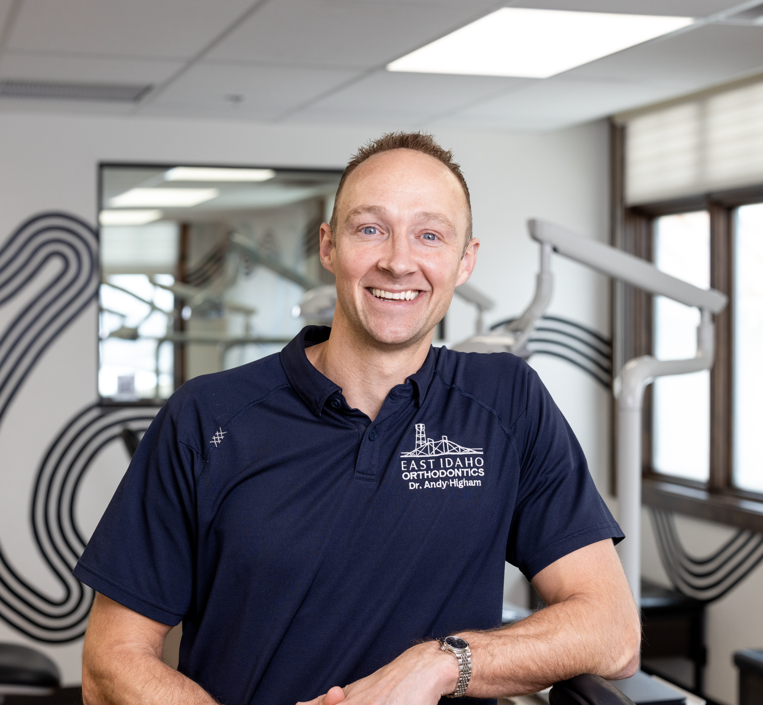 A smiling man wearing a navy polo shirt with 'East Idaho Orthodontics Dr. Andy Higham' logo, standing in a dental clinic with orthodontic equipment and window with blinds in the background.