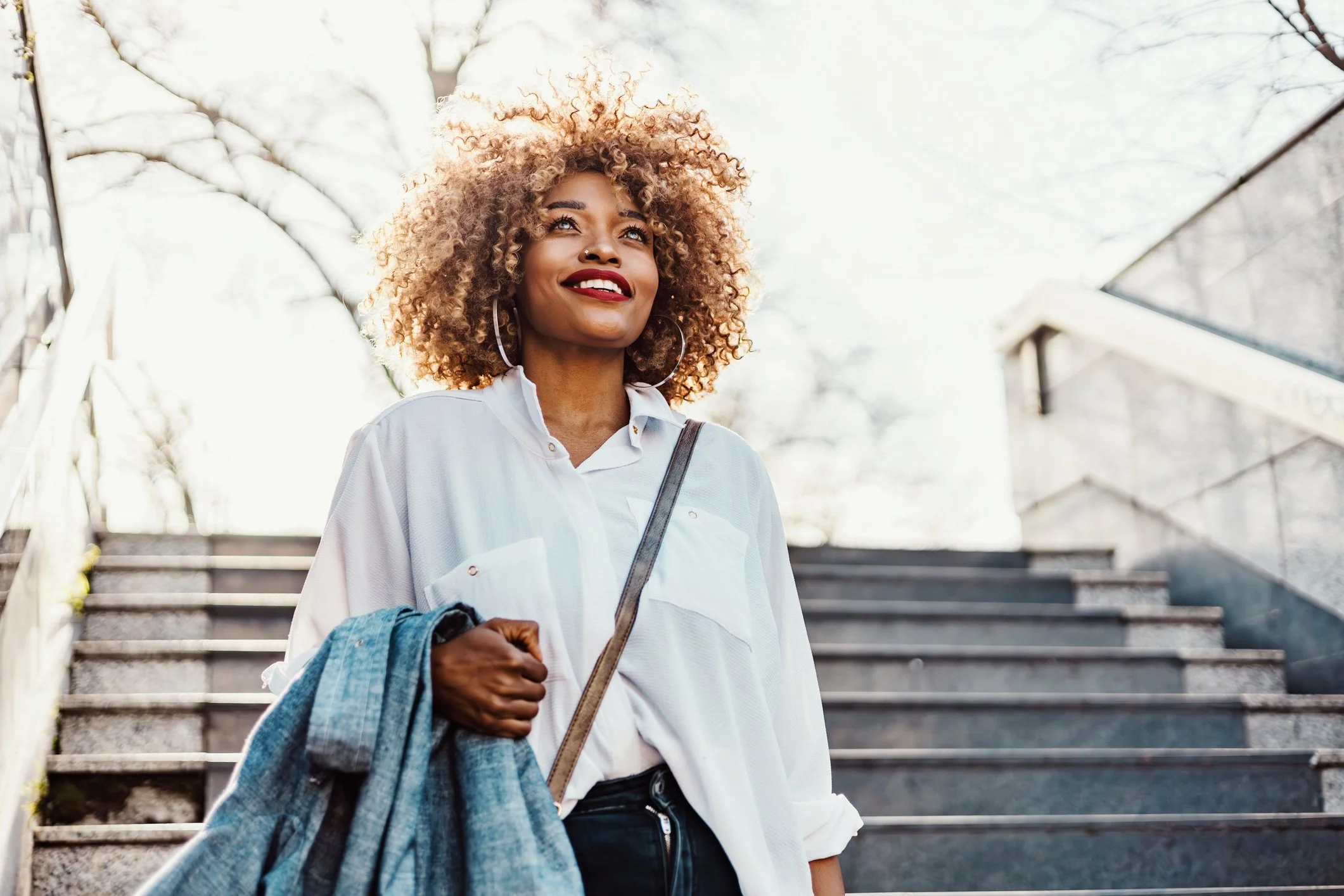 A woman with curly blonde hair, red lipstick, and hoop earrings smiling while holding a denim jacket, standing on outdoor stairs with a metal railing and trees in the background.