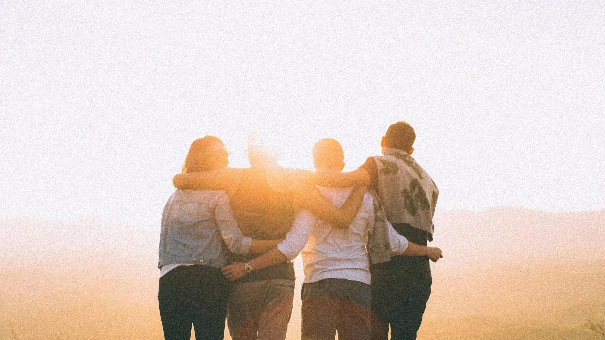 Four friends sitting together at sunset, with their arms around each other's shoulders, enjoying the view.
