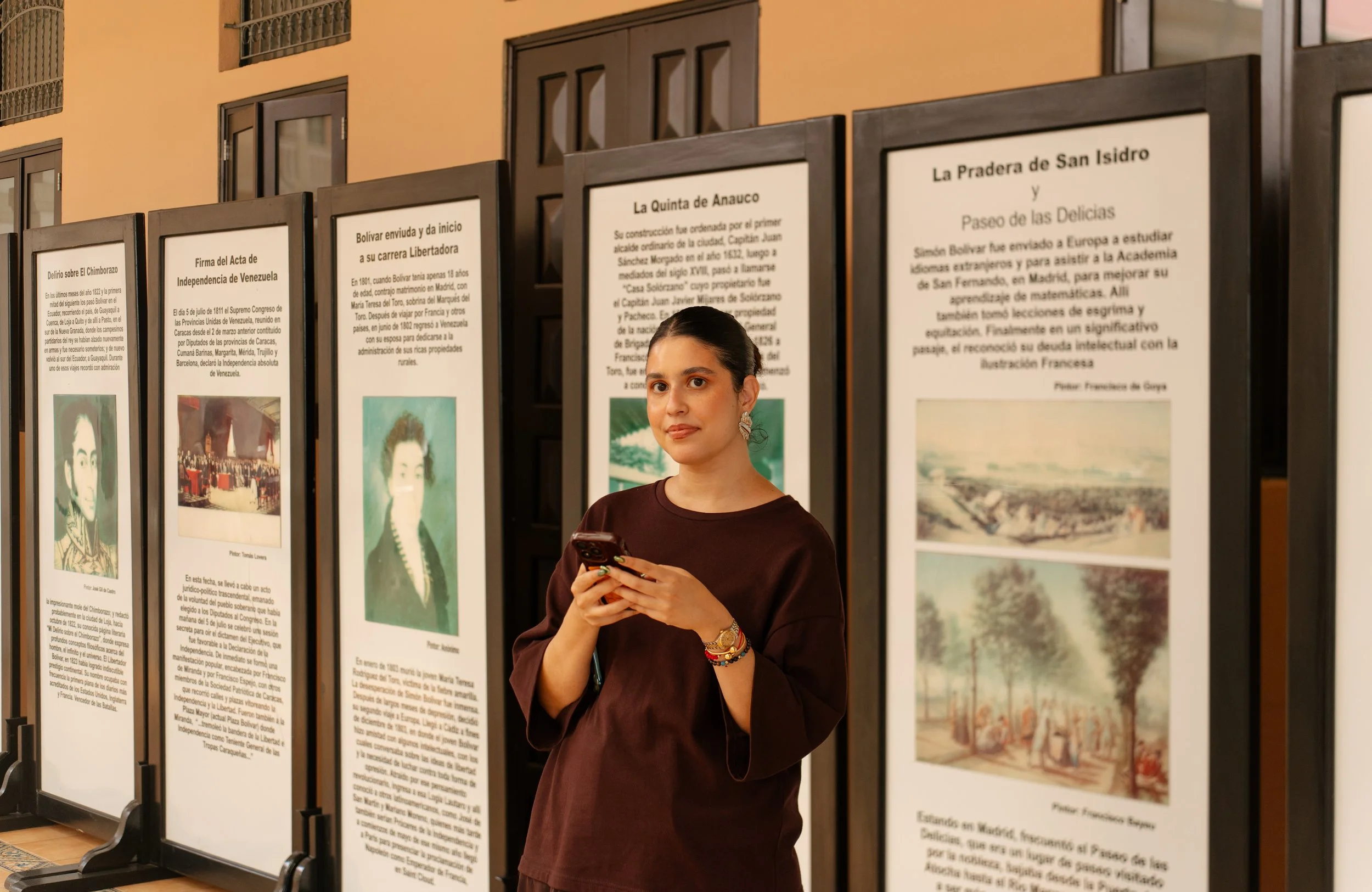 A young woman with dark hair in a bun, wearing a brown shirt, holding a smartphone, standing in front of a series of informational display panels at an indoor exhibition.