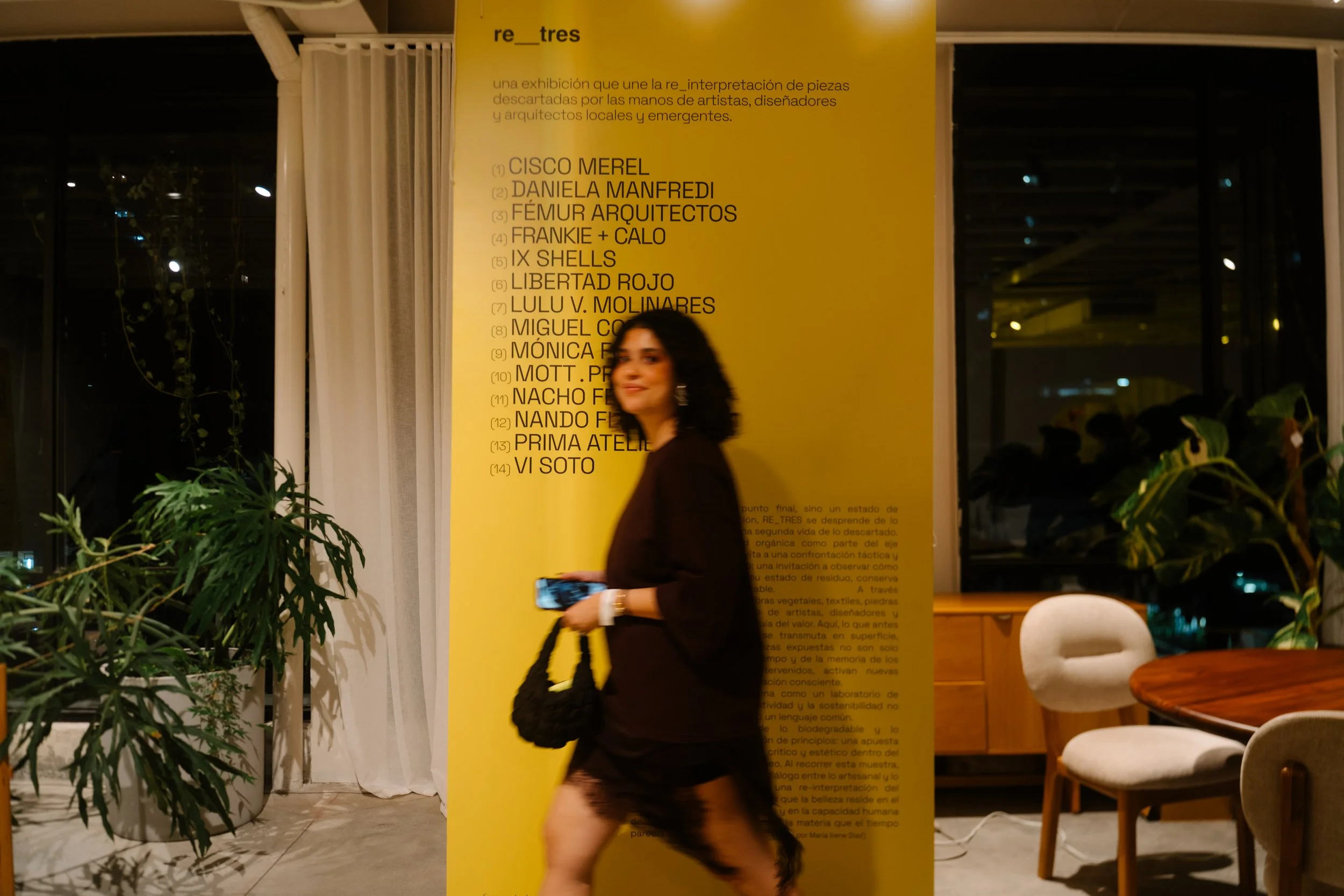 A woman with dark, curly hair, wearing a black dress with lace at the hem, walking past a yellow exhibition poster in a room with large windows and potted green plants.