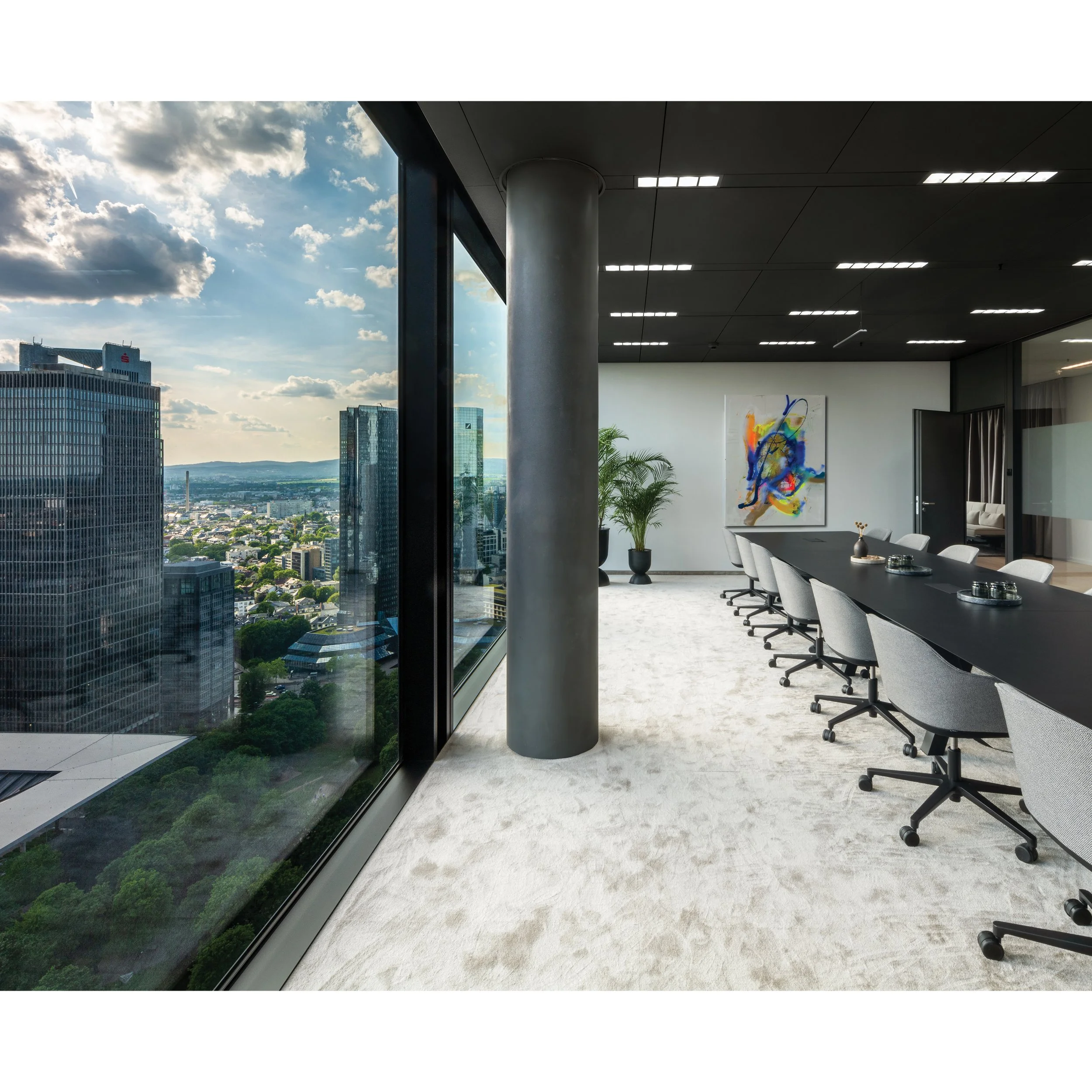 Modern office space with large glass window showing cityscape and sky, white carpeted floor, gray chairs around a table, colorful abstract painting on white wall, and a potted plant.