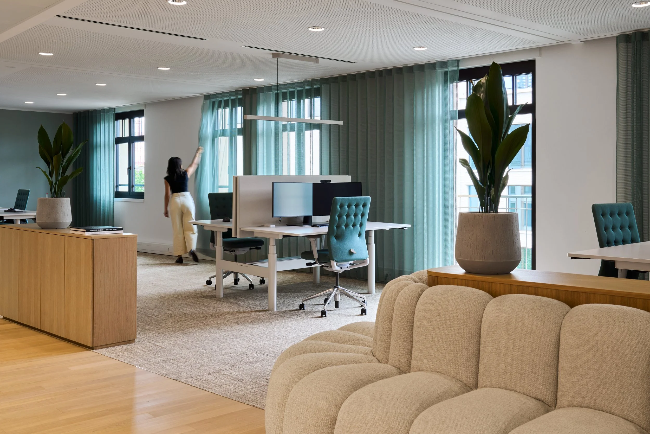 Modern gestaltete Bürofläche mit großen Fenstern, grünen Vorhängen und individueller Pflanzenauswahl für das Büro. Es sind Designer Schreibtische, komfortabel Stühle und Monitore ausgewählt worden