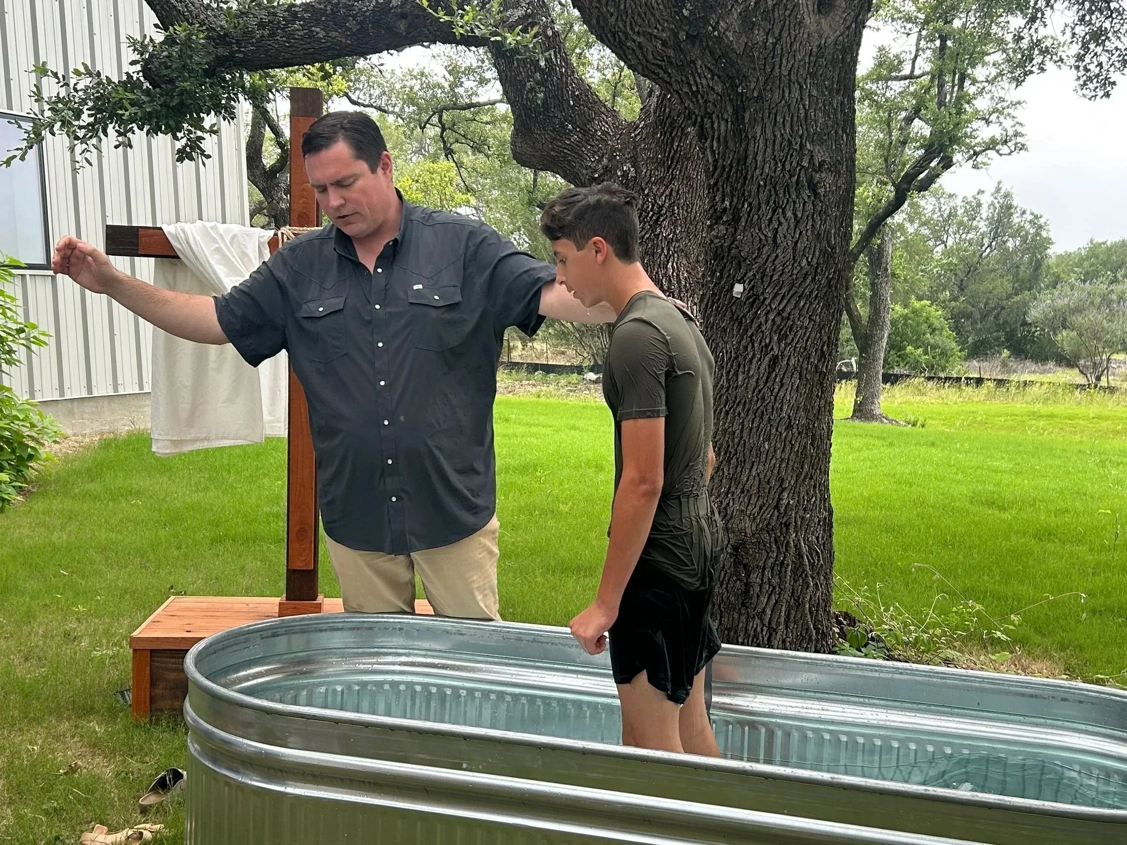 Two people, an adult and a teenager, are outdoors near a large tree with a thick trunk. They are engaging in an activity involving a metal tub, possibly some sort of ritual or tradition. The adult holds a cloth or towel, and the teenager stands in the tub, with water visible inside. Behind them, there is a wooden cross with a white cloth draped over it, and a building with metal siding is visible to the left. The scene takes place in a grassy area with additional trees and a fence in the background.