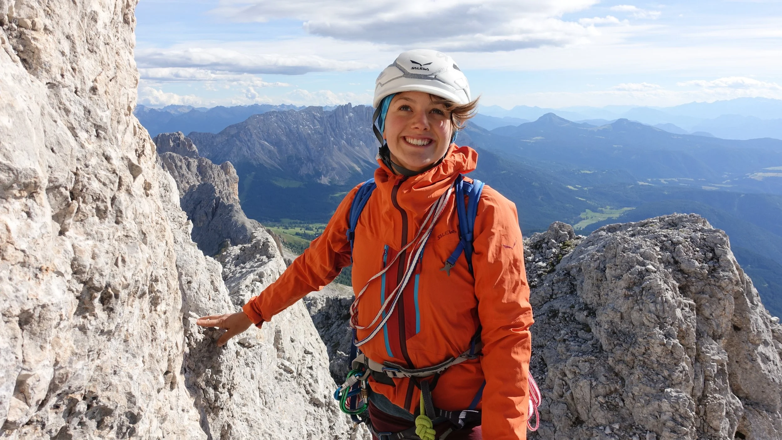 Lächelnde Bergsteigerin in orange Rucksack und Kletterhelm auf einer Bergspitze mit Blick auf die Bergkette im Hintergrund.