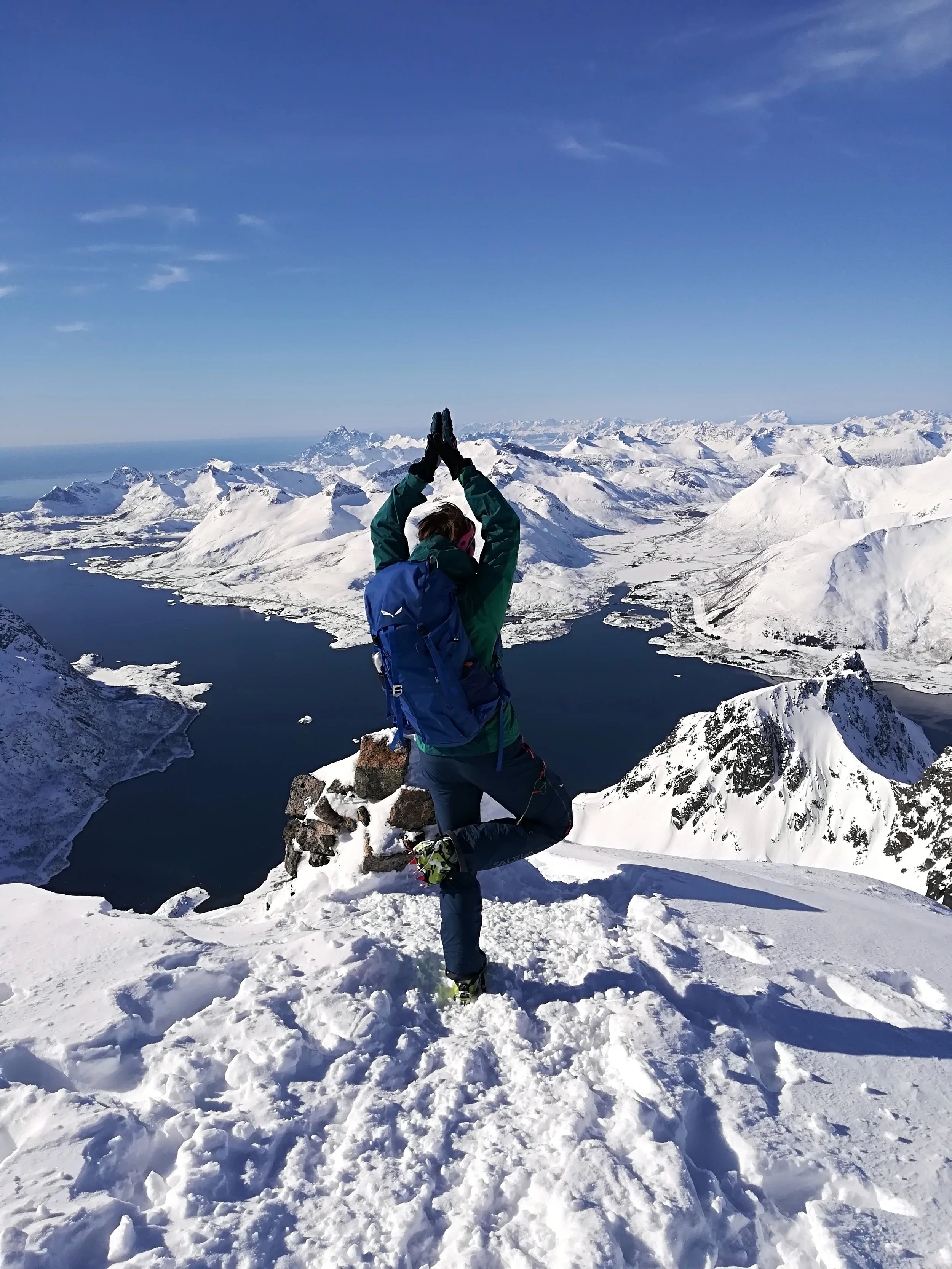 Eine Frau Winterkleidung steht auf einem schneebedeckten Berg in den Lofoten und macht eine Yoga-Pose mit den Händen über dem Kopf vor einer Landschaft mit verschneiten Bergen und dem Meer.