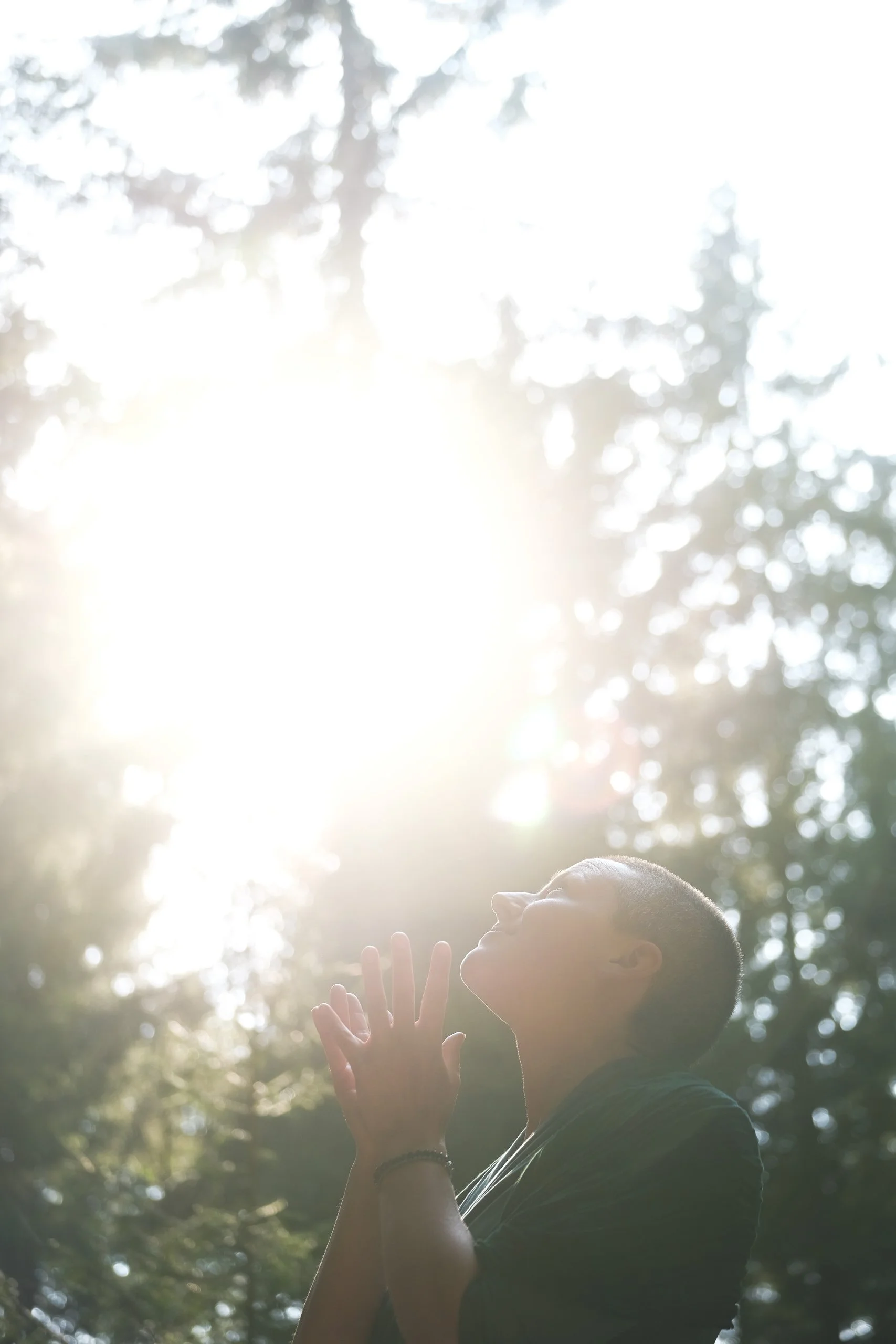 A person with closed eyes blankly facing the sun in a forested area with sunlight filtering through trees.