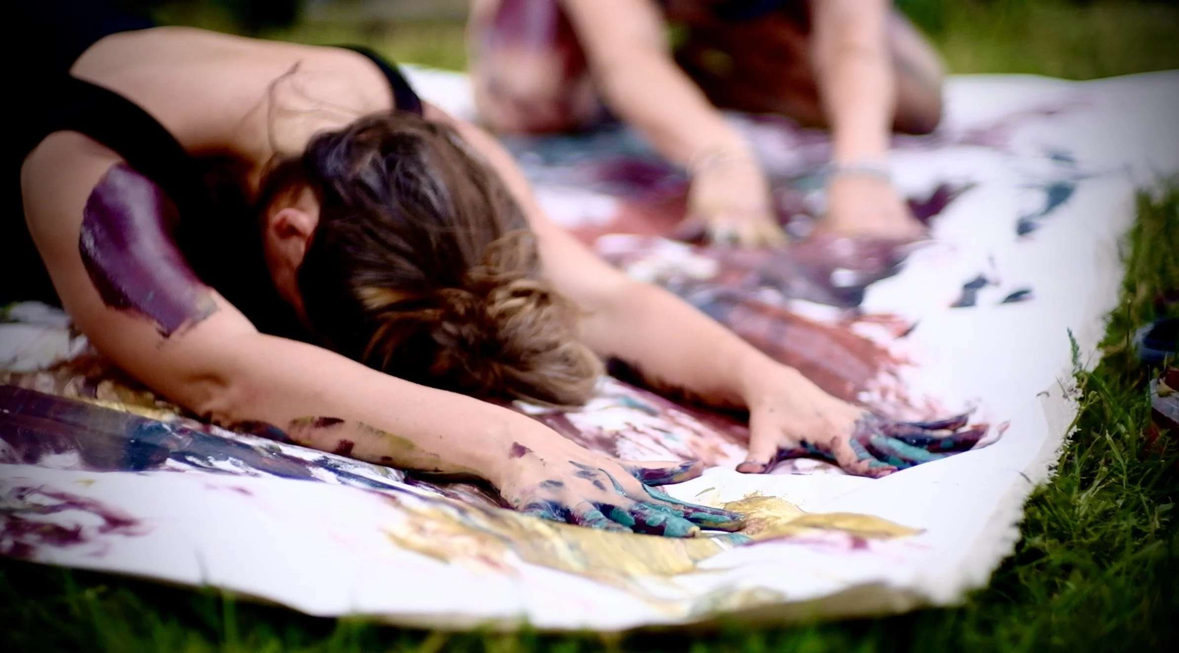 Zwei Frauen bemalen mit ihrem Körper mit bunten Farben ein großes Tuch, umgeben von Gras. Body-Art