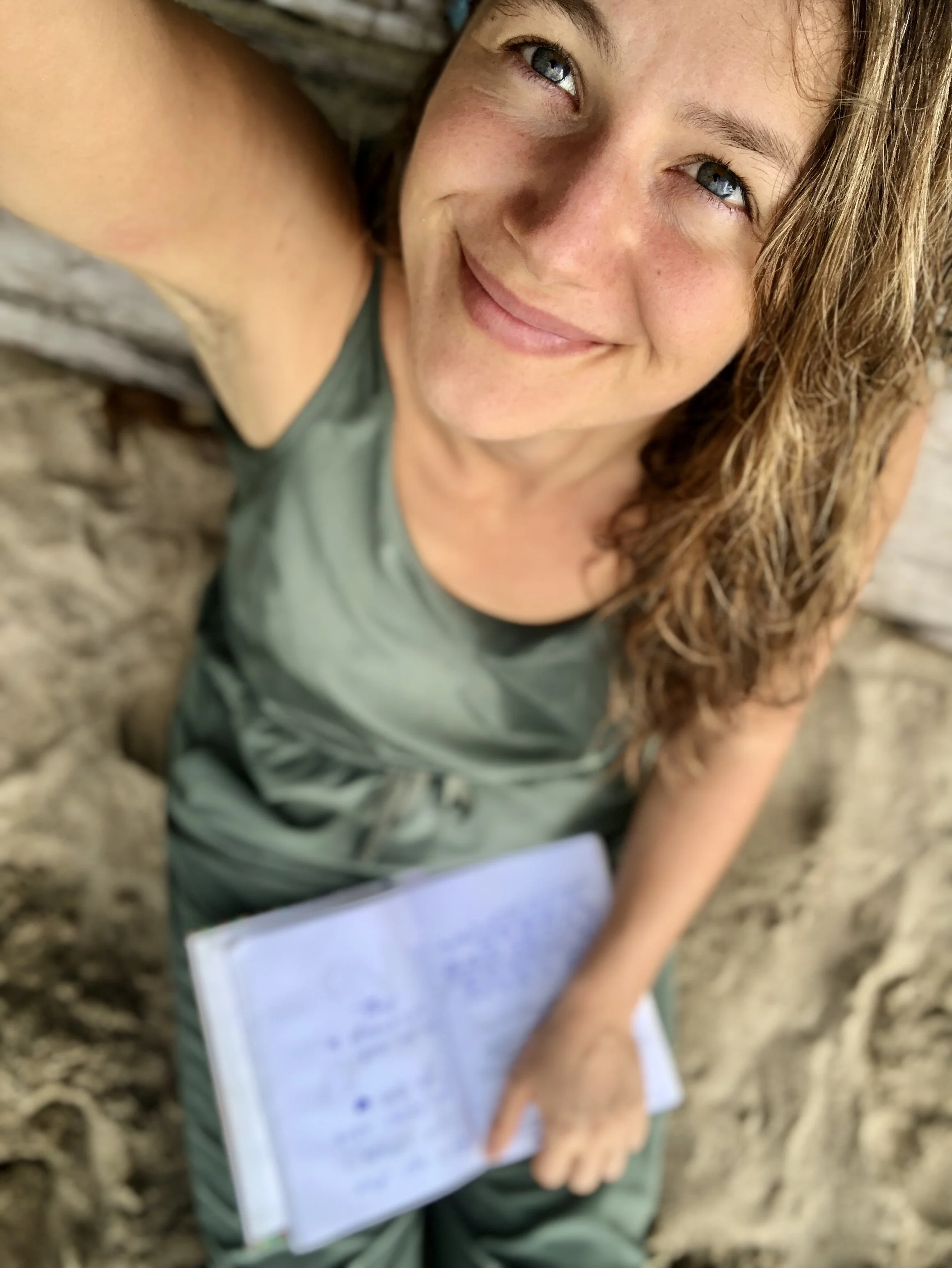 Junge Frau mit braunen, gewellten Haaren und blauen Augen, die auf dem Boden sitzt, ein Notizbuch in der Hand hält und lächelt.