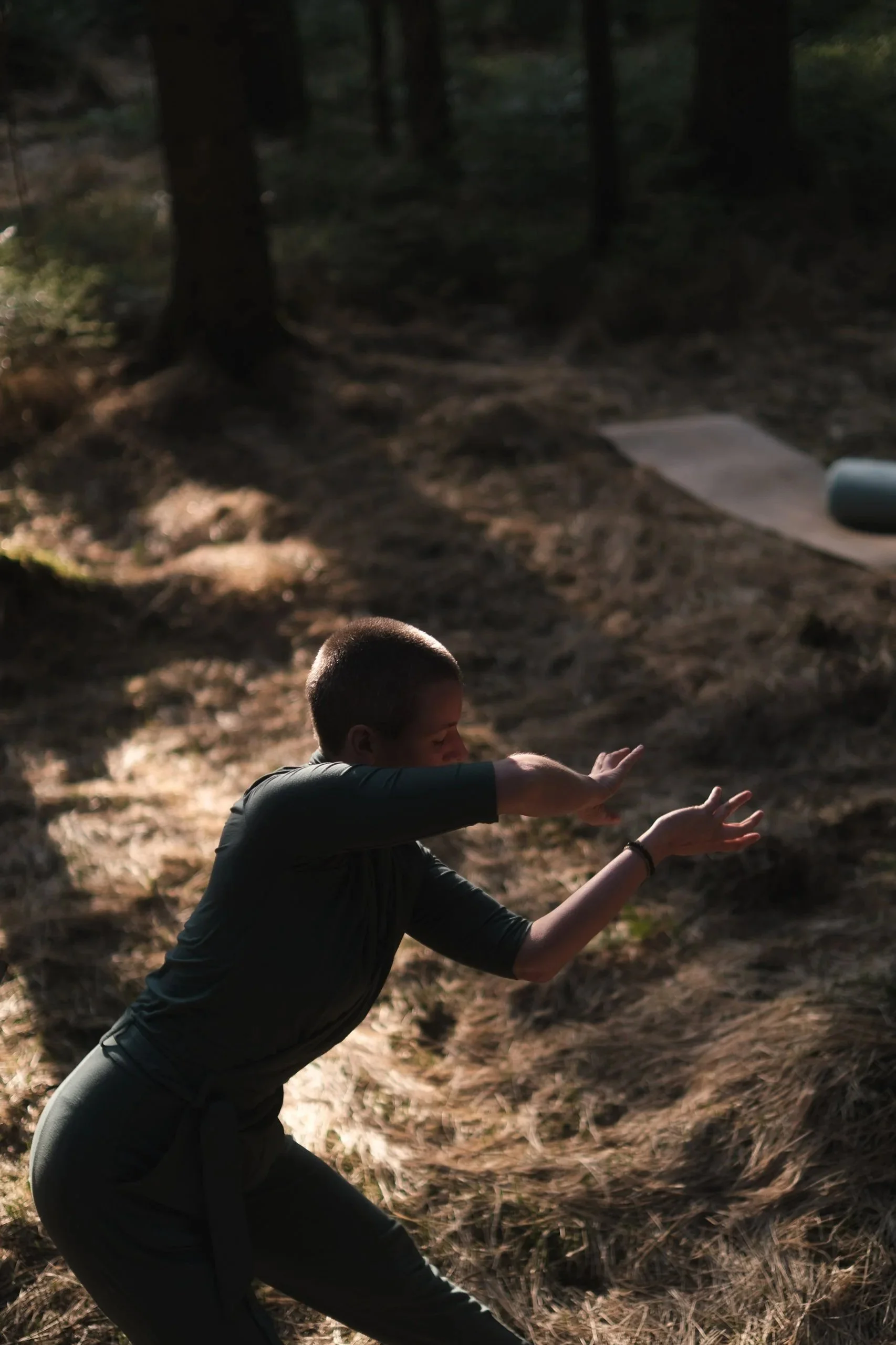 A person practicing yoga outdoors in a forest, kneeling on the ground with arms extended forward and palms facing down.