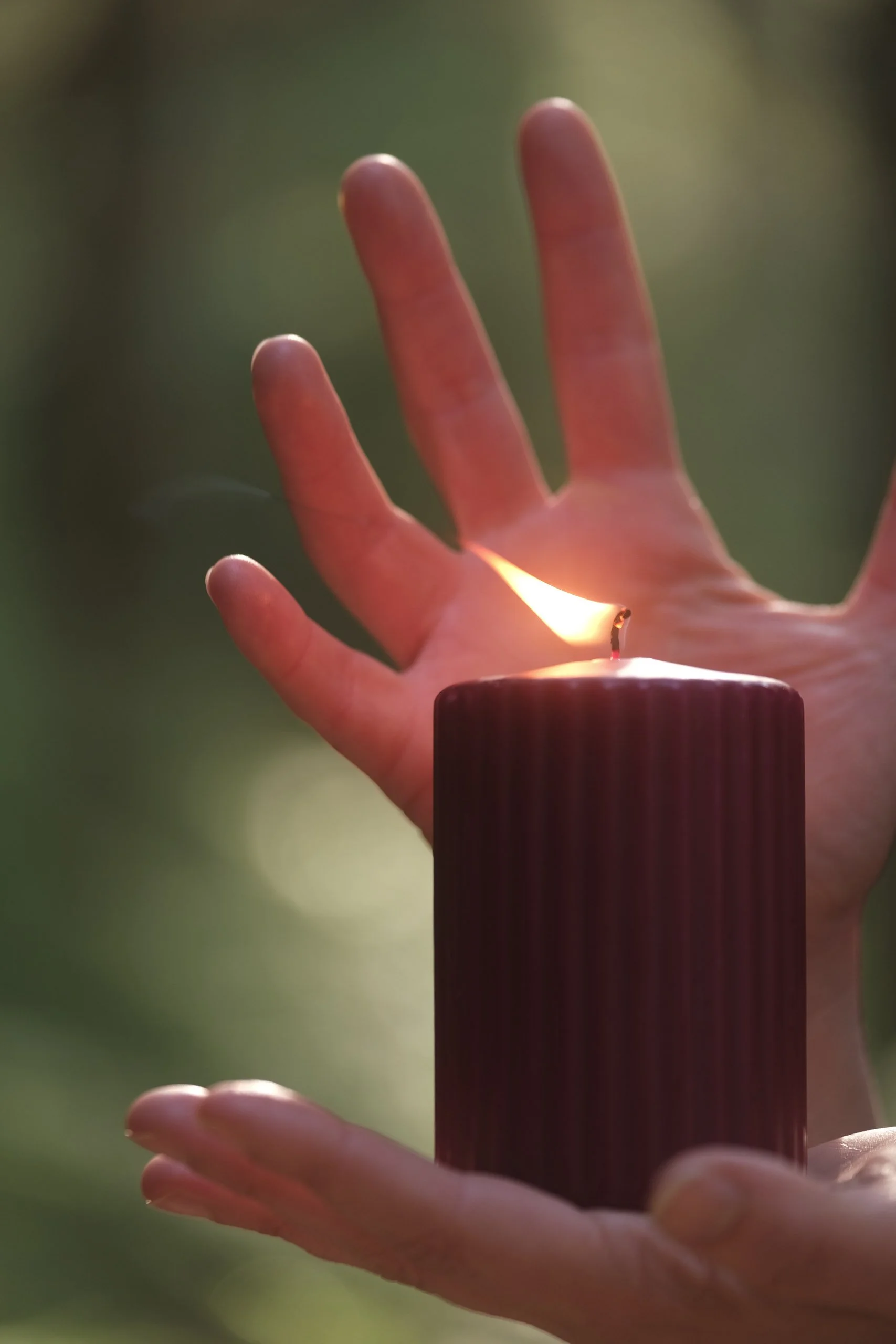 A hand delicately holding a candle with a small flame, in a dark wooden holder, against a blurred green background.