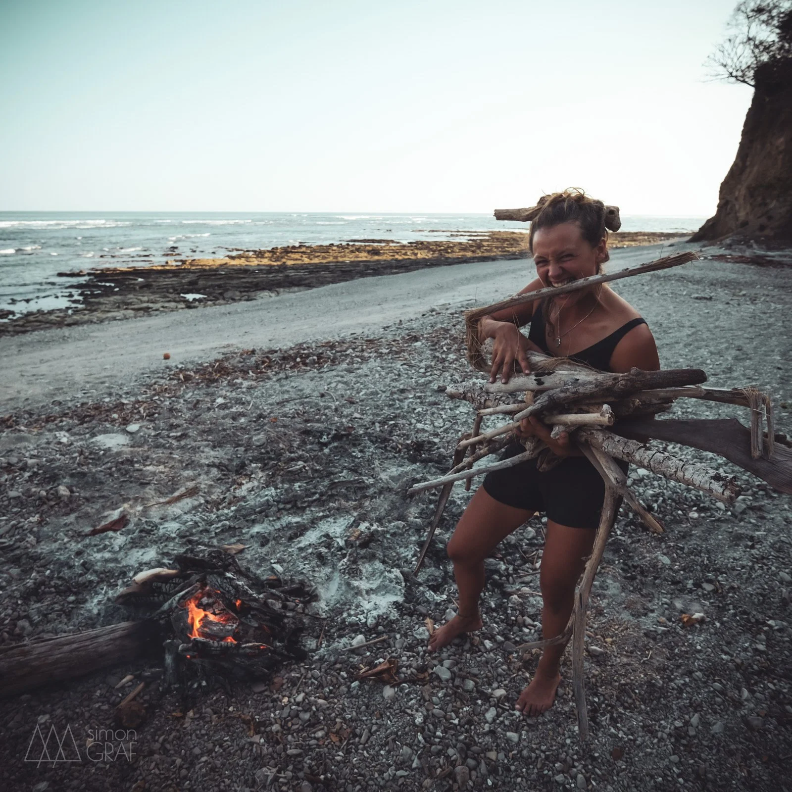 Eine Frau am Strand, die Holzstücke trägt, bei einem Lagerfeuer, mit Klippen im Hintergrund.