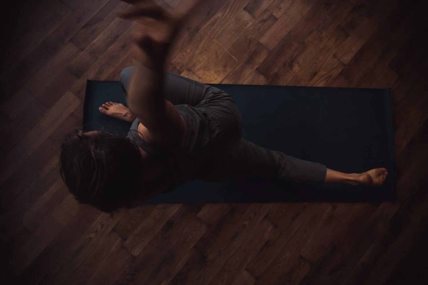 Frau macht Yoga auf einer Matte auf Holzboden, obere Perspektive, dunkle Kleidung, entspannte Atmosphäre.