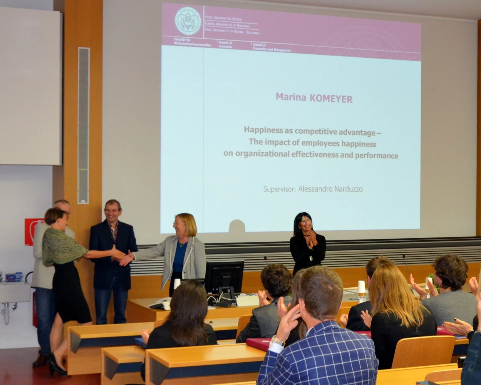A university presentation room with a large screen displaying a slide. The slide contains the logo and name of the Free University of Bozen-Bolzano, and the title "Happiness as competitive advantage — The impact of employees happiness on organizational effectiveness and performance" by Marina Komeyer, supervised by Alessandro Narduzzo. In front of the screen, four people are standing; three are shaking hands, and one woman is clapping while smiling. The audience is seated, clapping and looking at the speakers.