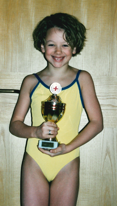 A young girl with curly hair in a yellow and blue swimsuit holding a trophy with a medical cross symbol.