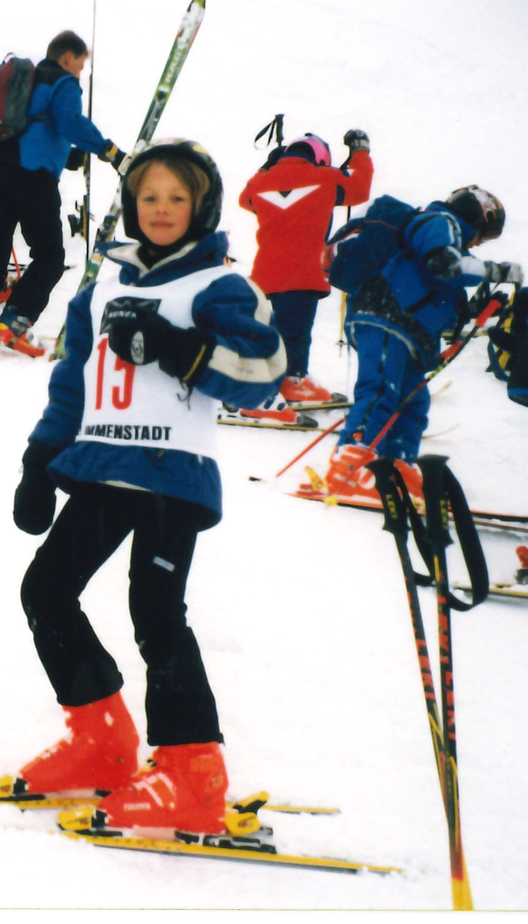 Children skiing on a snowy mountain, wearing helmets and ski gear, with some standing and others adjusting their skis.