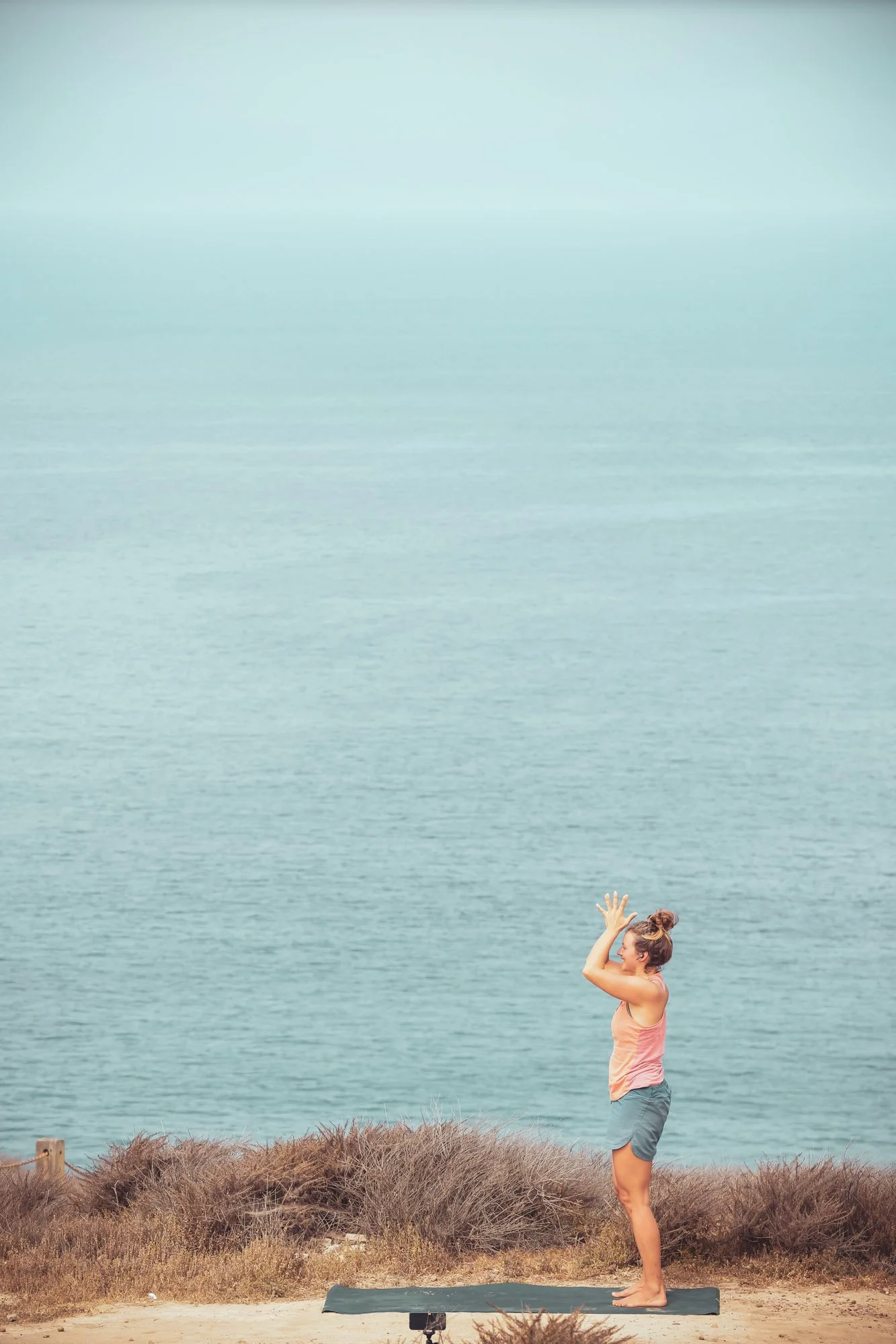 Frau macht Yoga im Freien auf einer Klippe mit Blick auf den Ozean, trägt Sportkleidung und steht auf einer Yogamatte.