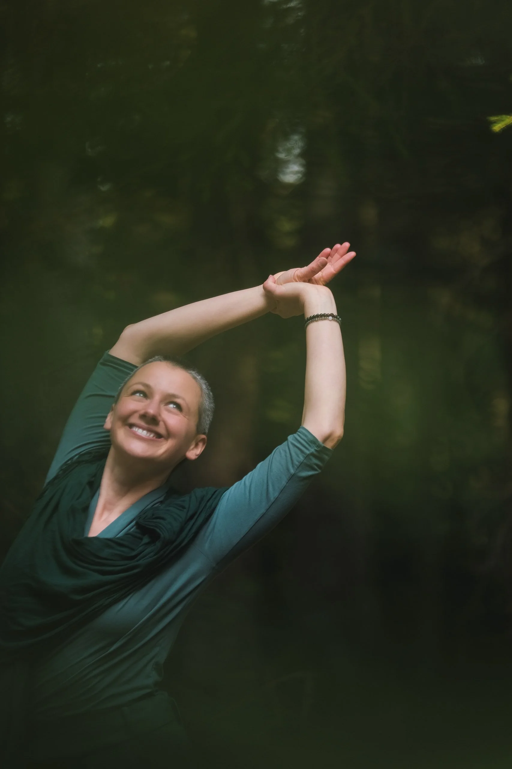 Eine lachende Frau mit kurzer grauer Frisur, die im Grünen steht und eine Hand in der Luft hält, während sie in den Himmel zeigt.