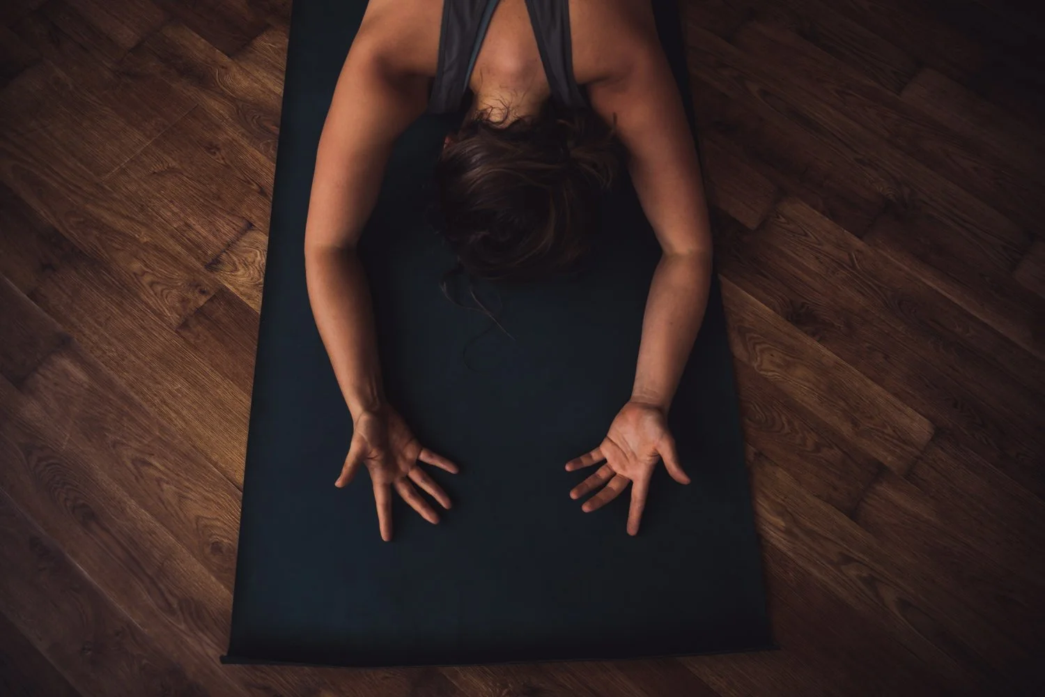 Frau macht Yoga auf einer schwarzen Yogamatte auf Holzfußboden, mit ausgestreckten Armen, Blick nach unten, Dunkelraum mit warmem Licht.