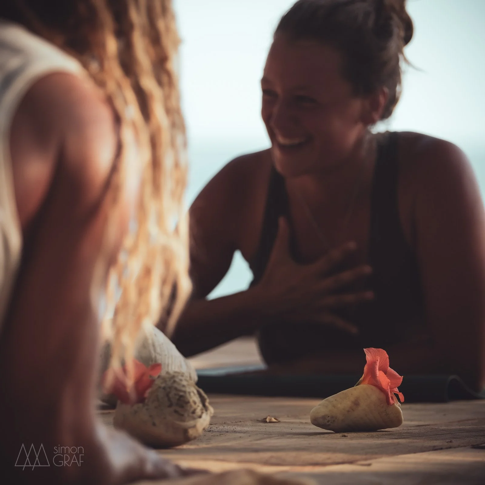 Zwei Frauen lachen sich beim Yoga an während ihre Hand auf dem Herzen ruht.