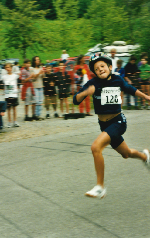 Young boy wearing a helmet, running a race with a bib number 120, with spectators cheering behind a yellow barrier on a cloudy day.