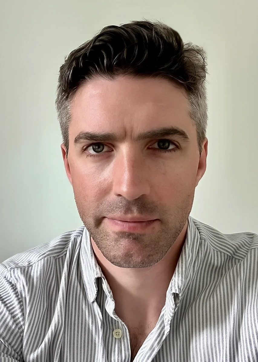 A close-up photo of a man with dark hair, blue eyes, and light facial stubble, wearing a striped button-up shirt with the collar open, standing against a plain light-colored background.