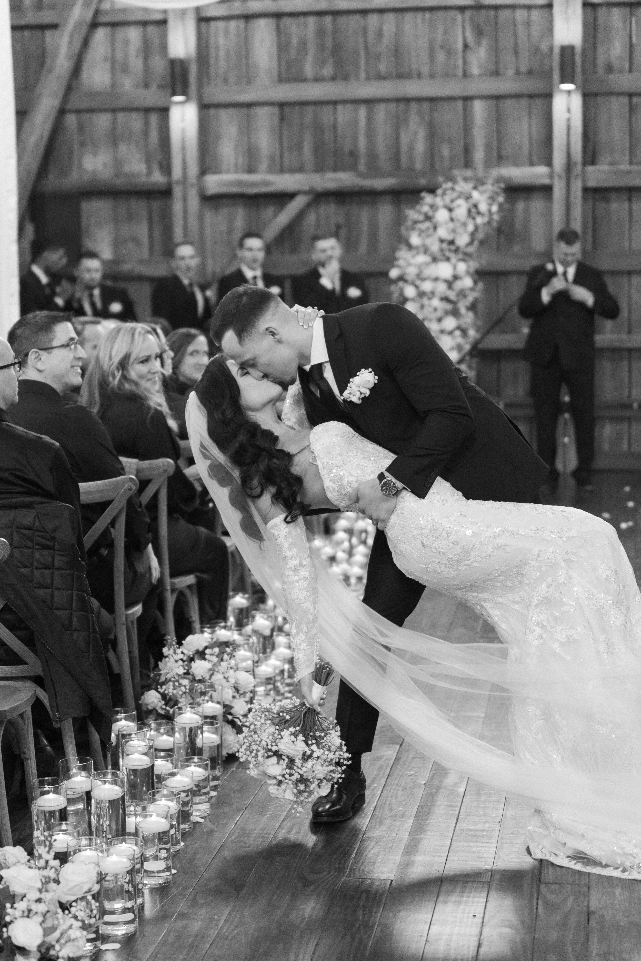 A black-and-white photo of a groom dipping his bride in front of guests during a wedding ceremony in a rustic wooden venue.