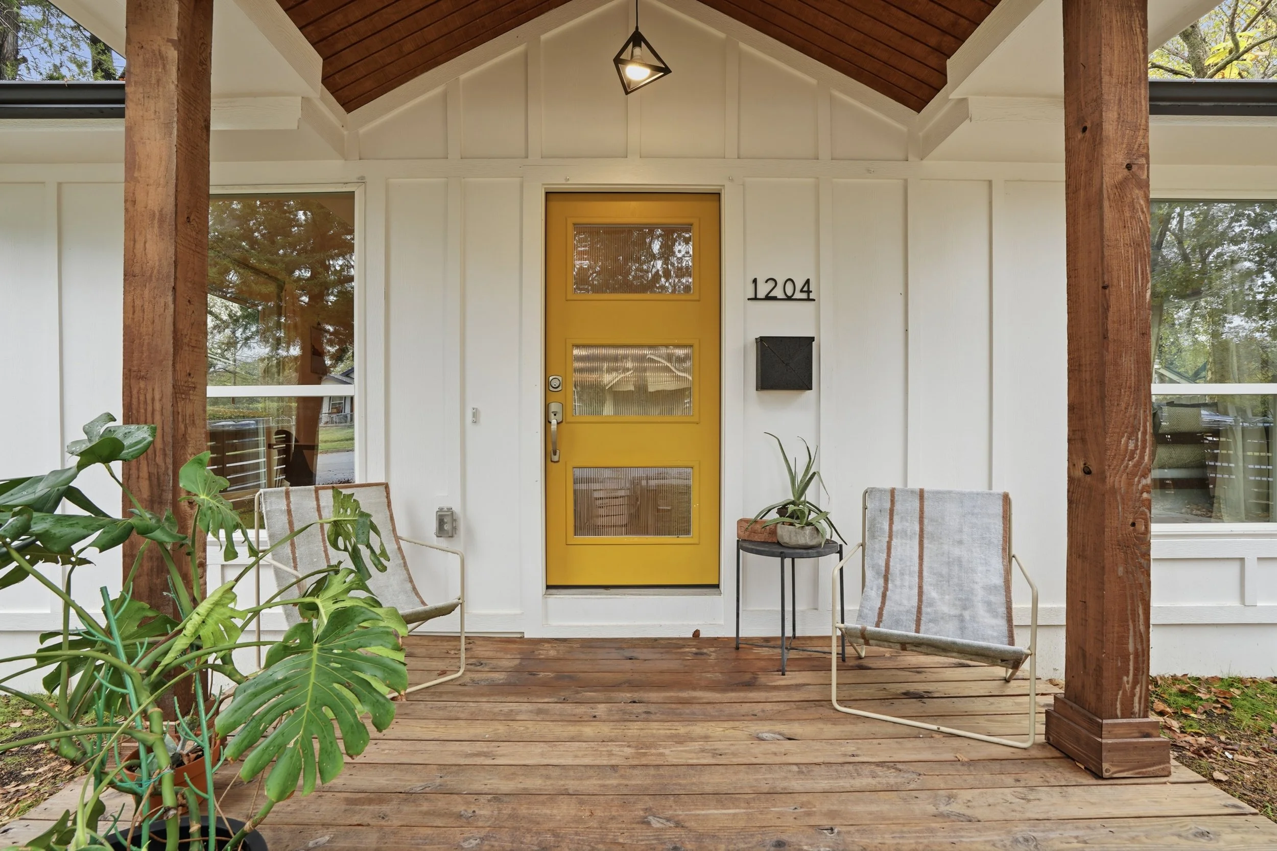 Front porch with yellow door, white siding, two chairs with striped cushions, potted plants, and wooden posts, under a wooden ceiling with a hanging light fixture.