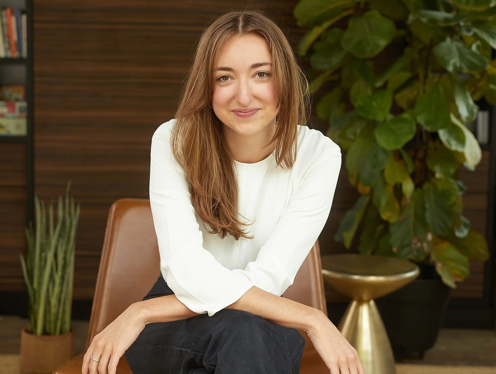 A woman with long, light brown hair sits on a brown chair, smiling at the camera, in a room with wooden paneling and large green plants.