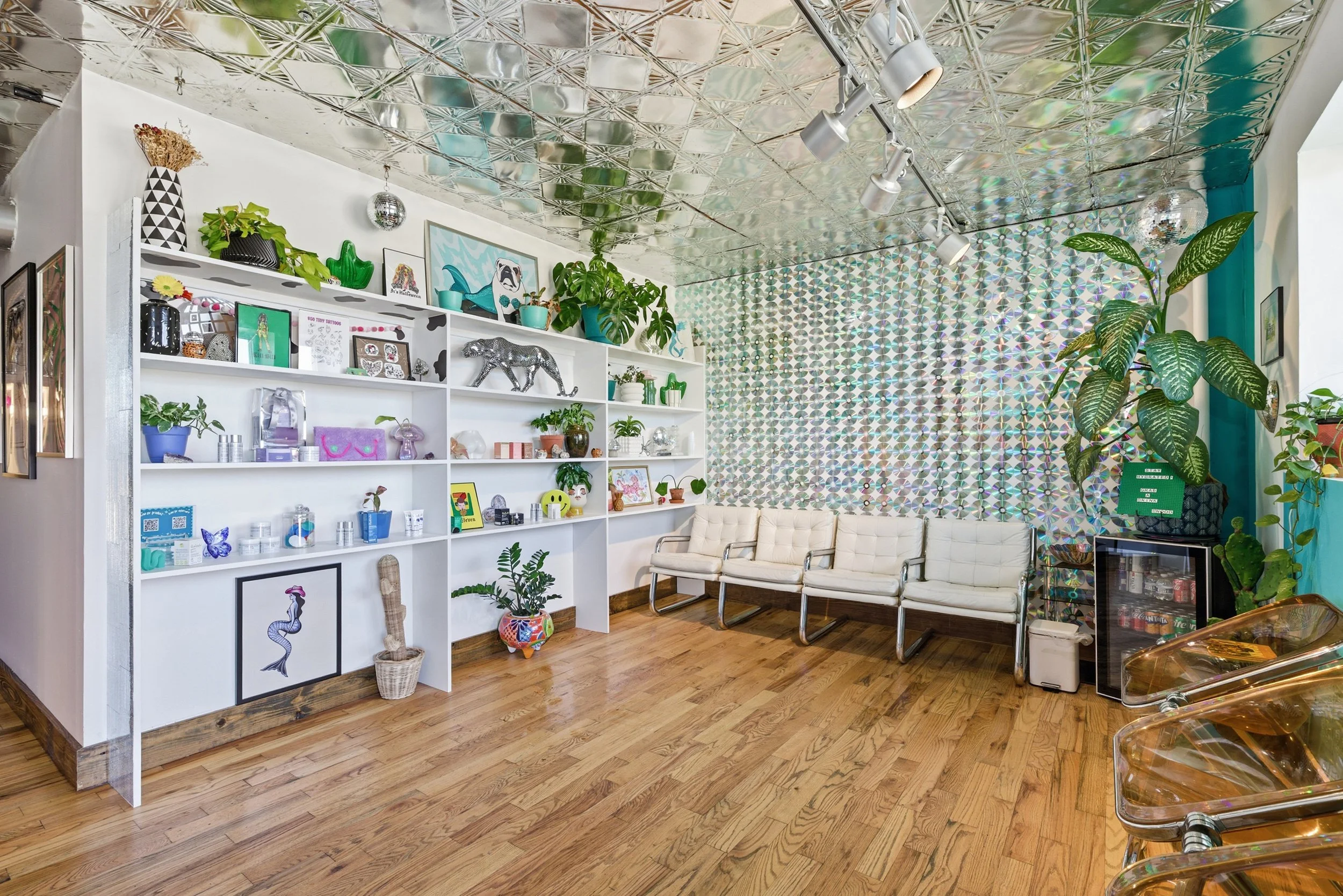 A brightly decorated room with a shiny metallic ceiling, white shelving filled with plants, artwork, and decorative objects, and white chairs along the wall. There are various plants, art pieces, and a disco ball hanging from the ceiling.