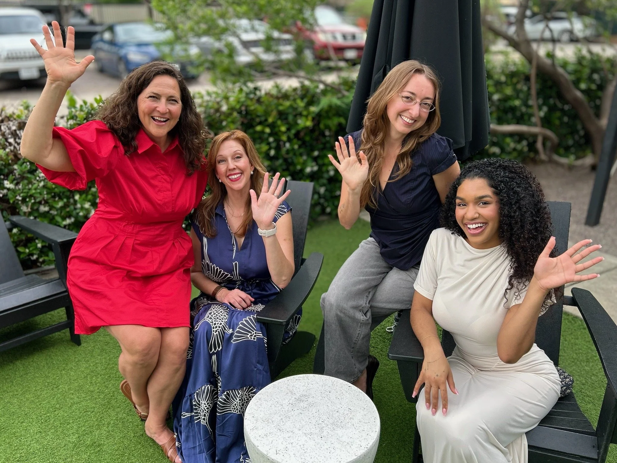 Four smiling women sitting and standing outdoors, waving at the camera, with green bushes and parked cars in the background.