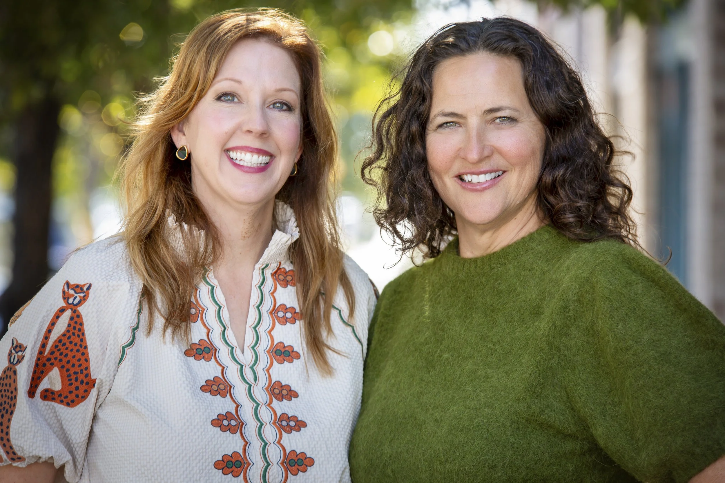 Two smiling women standing outdoors with trees in the background. One woman has red hair and is wearing a white embroidered blouse. The other woman has dark curly hair and is wearing a green sweater.