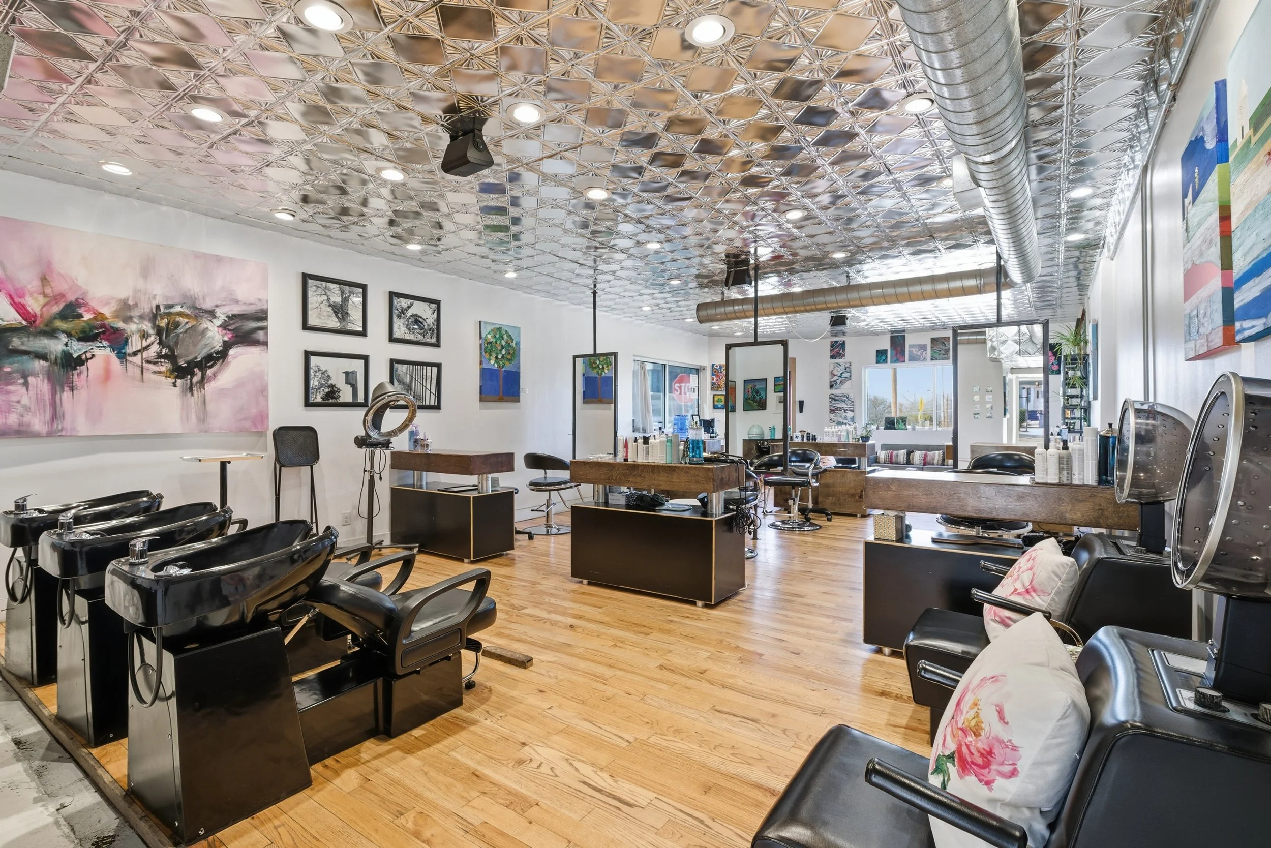 Interior of a hair salon with multiple styling chairs, mirrors, artwork on walls, and a metallic ceiling.