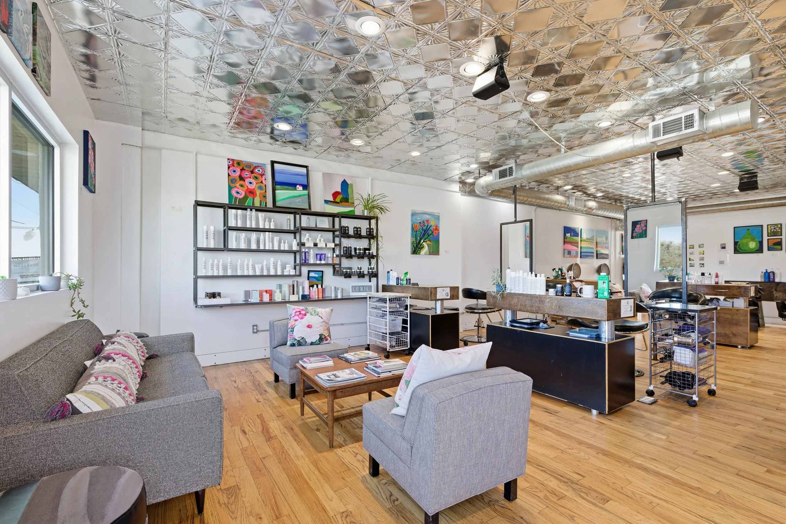A cozy hair salon with a leather sofa, a gray armchair with a floral pillow, and a wooden coffee table with magazines. Shelves with hair products, colorful artwork on the walls, and a large mirror behind the styling station. The ceiling has a shiny, 