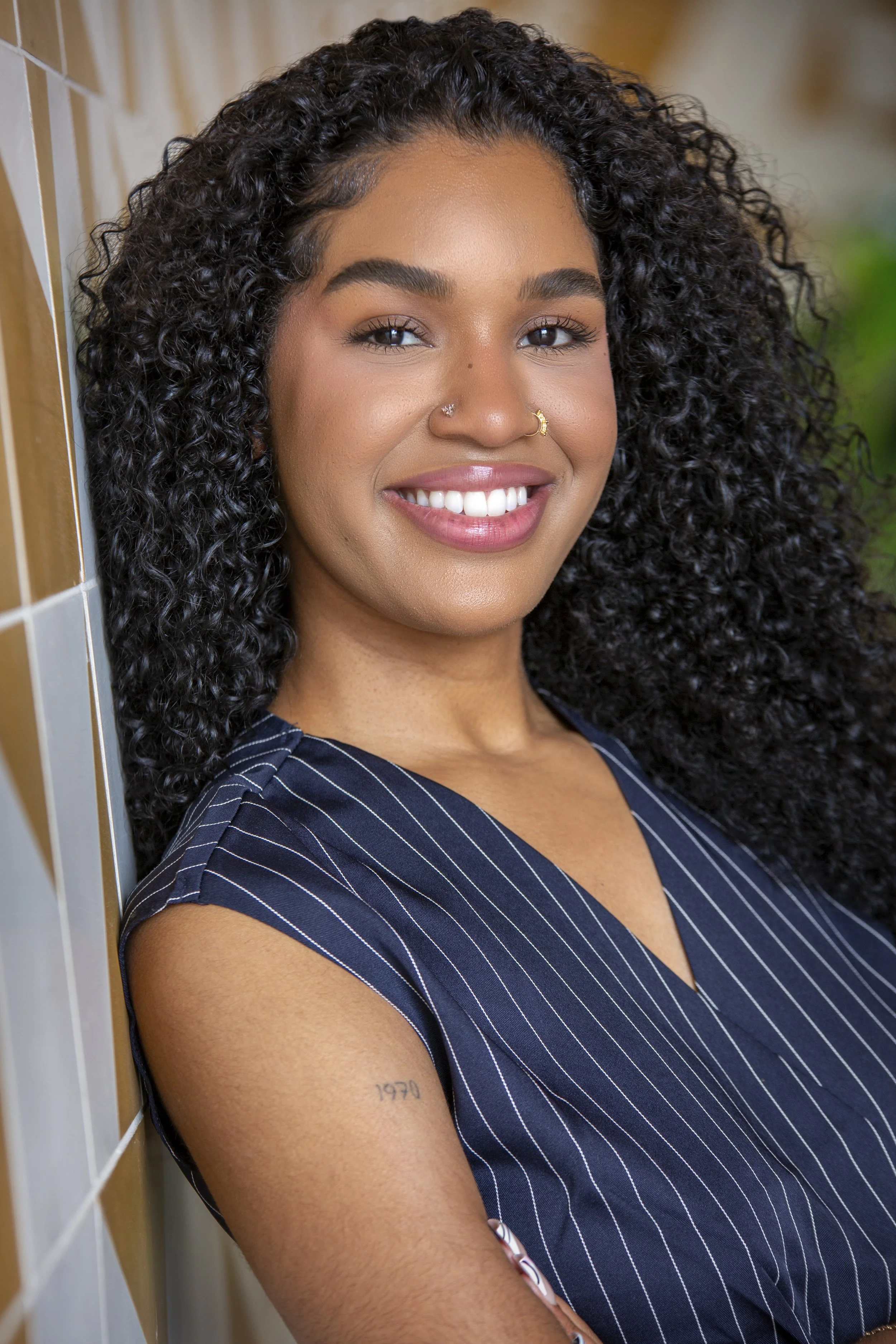 A woman with curly hair standing in a modern, well-lit living room, wearing a black suit with a white top and white heels.