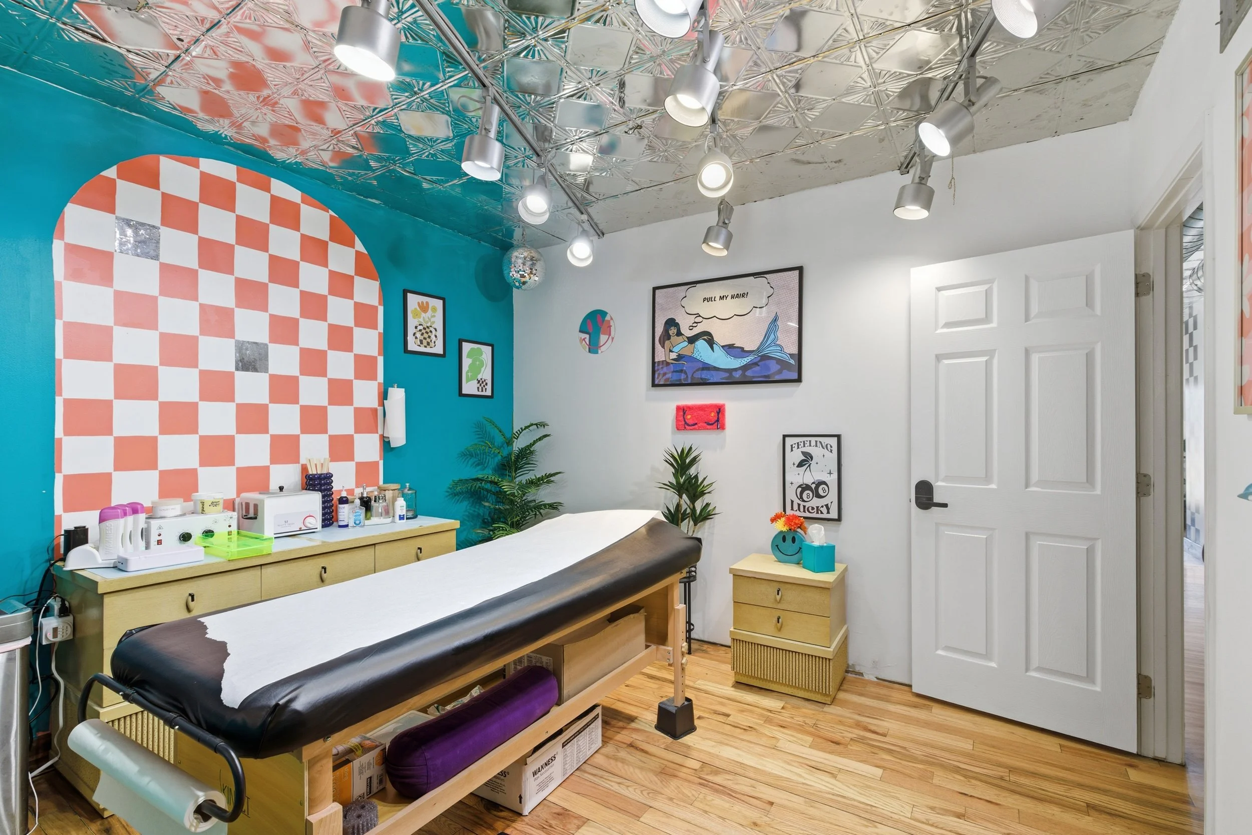 Medical examination room with a black examination table, a wooden cabinet with medical supplies, a white wall with colorful artwork including a sea creature and a mermaid, a blue and red checkered wall section, and a ceiling with metallic tiles and t