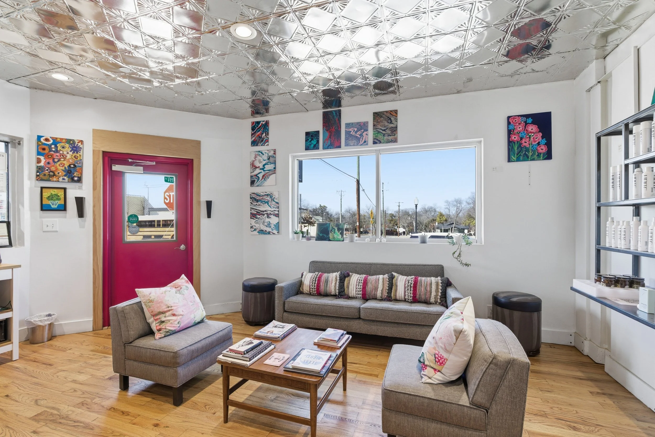 A cozy interior of an art gallery with white walls and a shiny, patterned metallic ceiling. Several colorful paintings are displayed on the walls. There is a large window letting in natural light, and a pink door with a small window. The room is furn