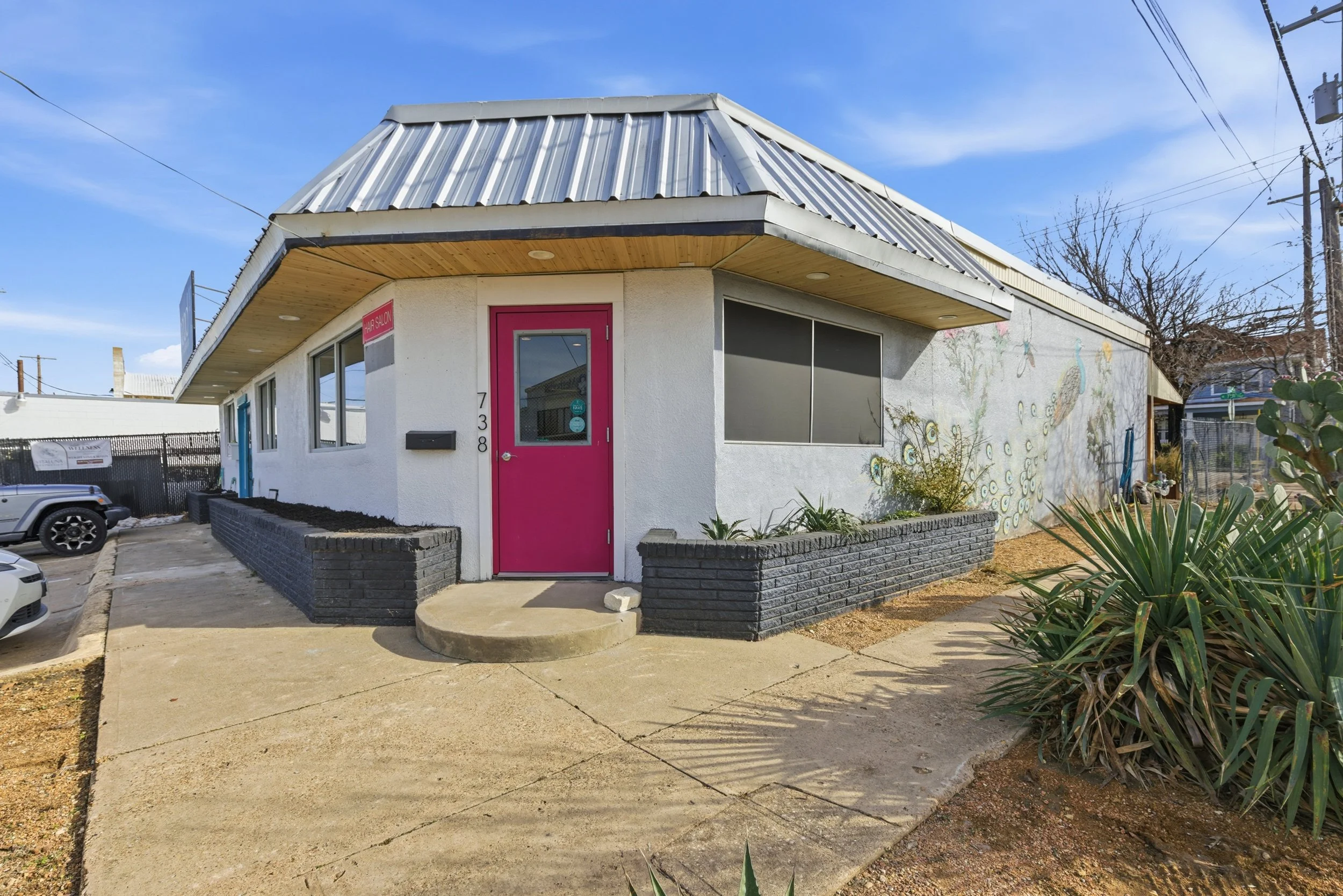 Building with white walls and a colorful pink door, featuring mural art of peacocks and flowers, with a small front porch and parking lot, under a blue sky.