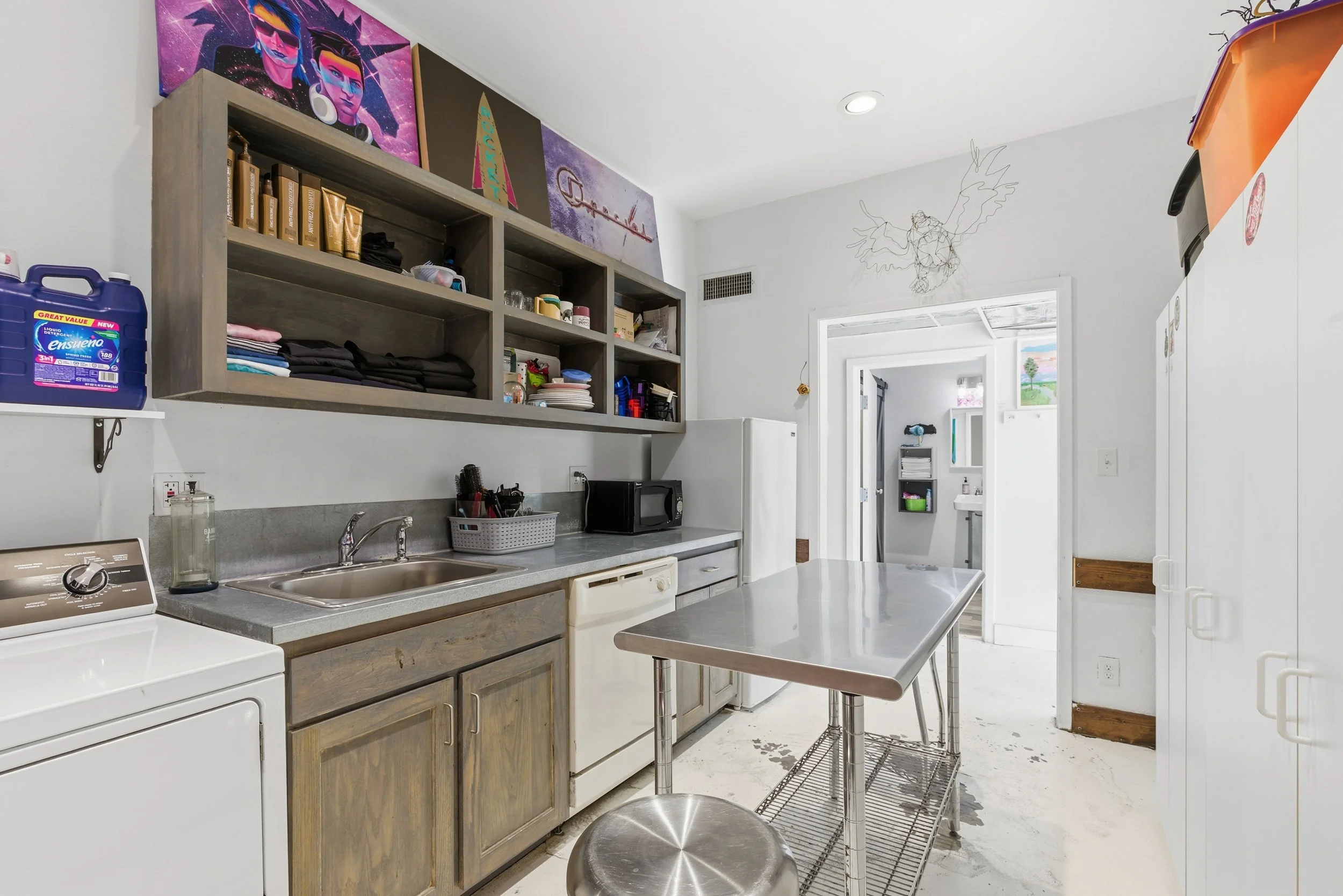 A kitchen with a white walls and ceiling, wooden cabinets, a gray countertop with a sink, microwave, and various kitchen items. Open shelves above hold dishes and small items. A small metal table with a black cushioned stool. Wall art, including a pa