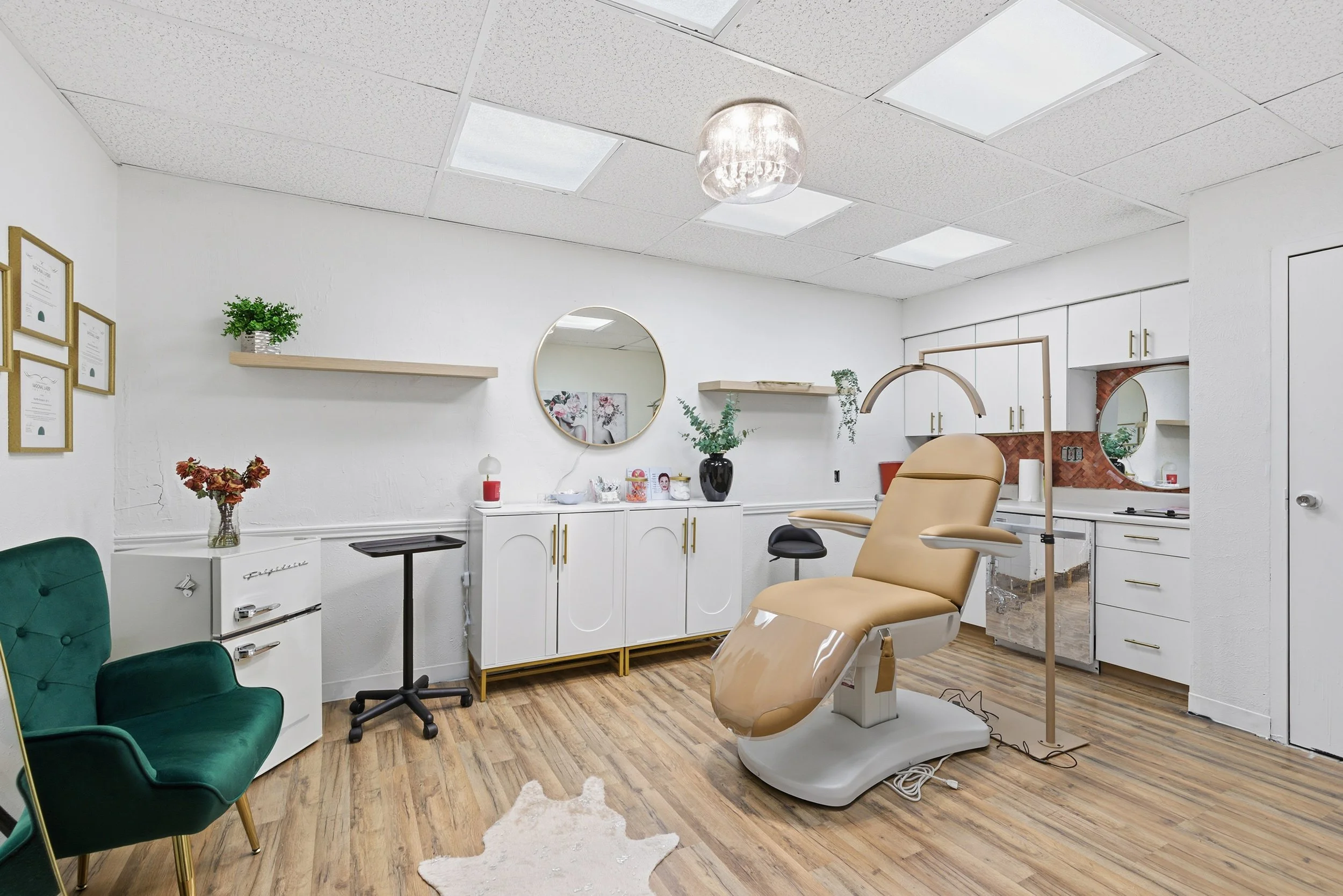A medical examination room with a dental chair, green chair, white cabinets, wall-mounted shelves, mirrors, plants, framed certificates, and ceiling lights.