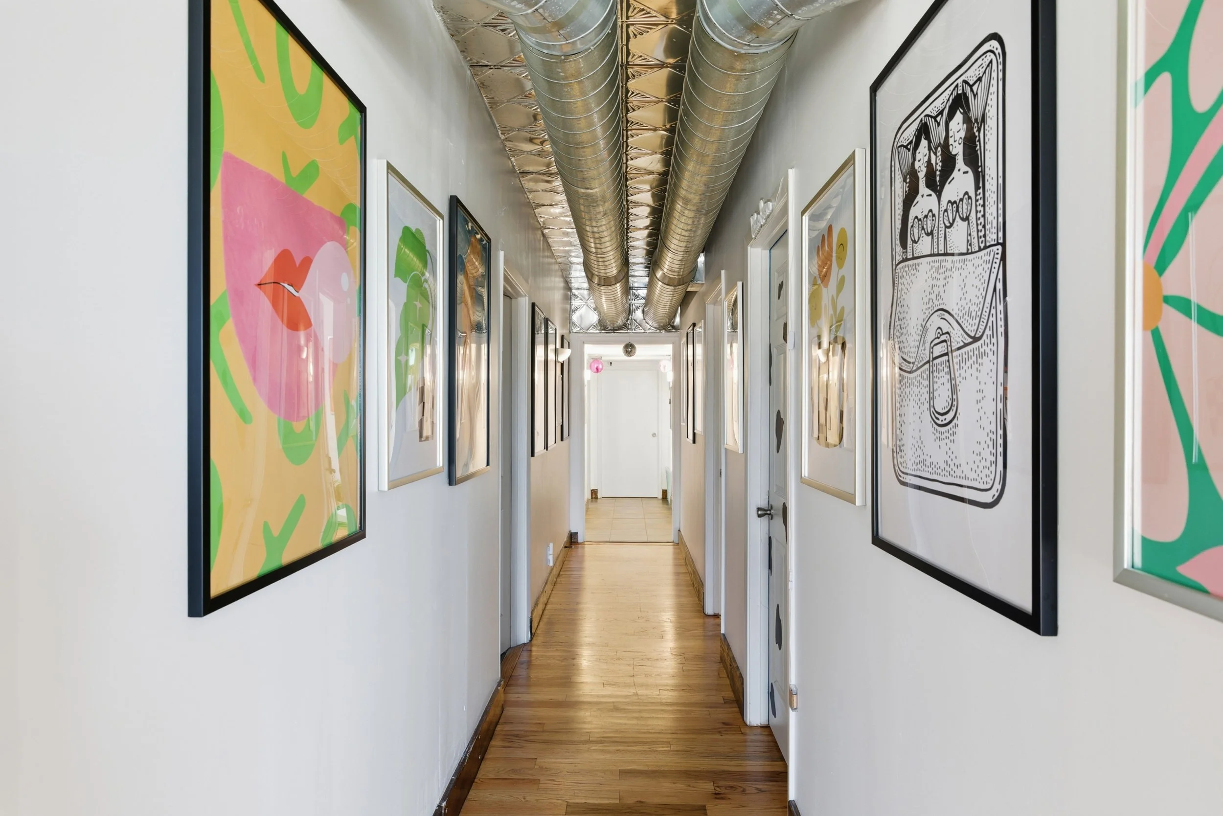Long hallway with wooden floors, white walls decorated with colorful and black-and-white framed artwork, and an exposed metallic ventilation duct running along the ceiling.