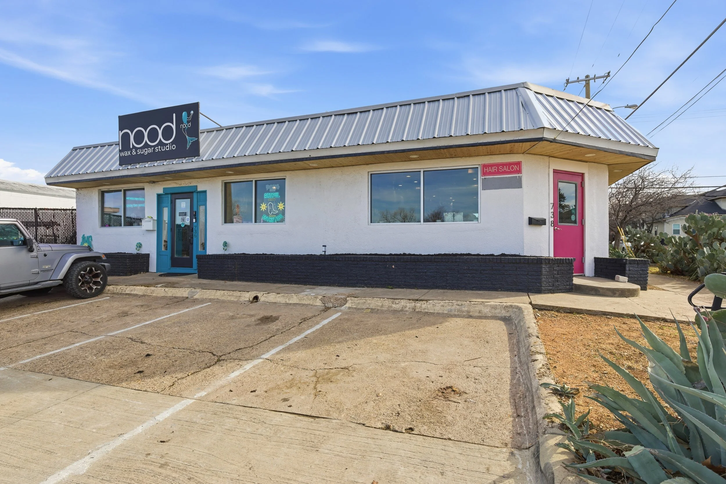 A single-story commercial building with a metal roof, white exterior walls, two large windows, a blue door, and a pink door. A parking lot with marked spaces and a parked gray Jeep in front. Signs for a wax and sugar studio and a hair salon. Dry land