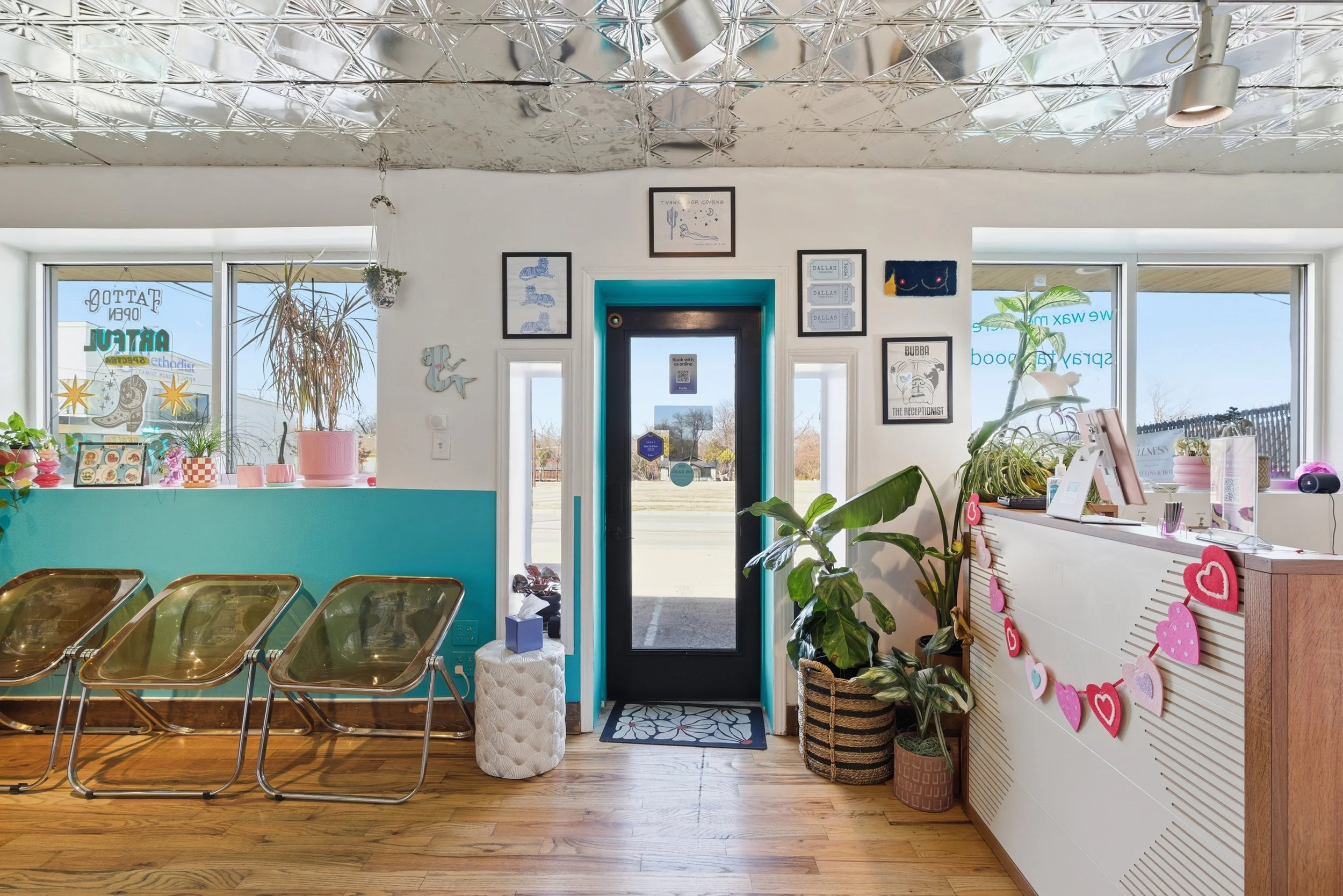 Interior of a bright cafe with wooden flooring, pink and teal decor, potted plants, wall art, and a row of gold chairs pressed against a teal counter. The front door is black with a window, and large windows outside show a sunny day with a fence and 